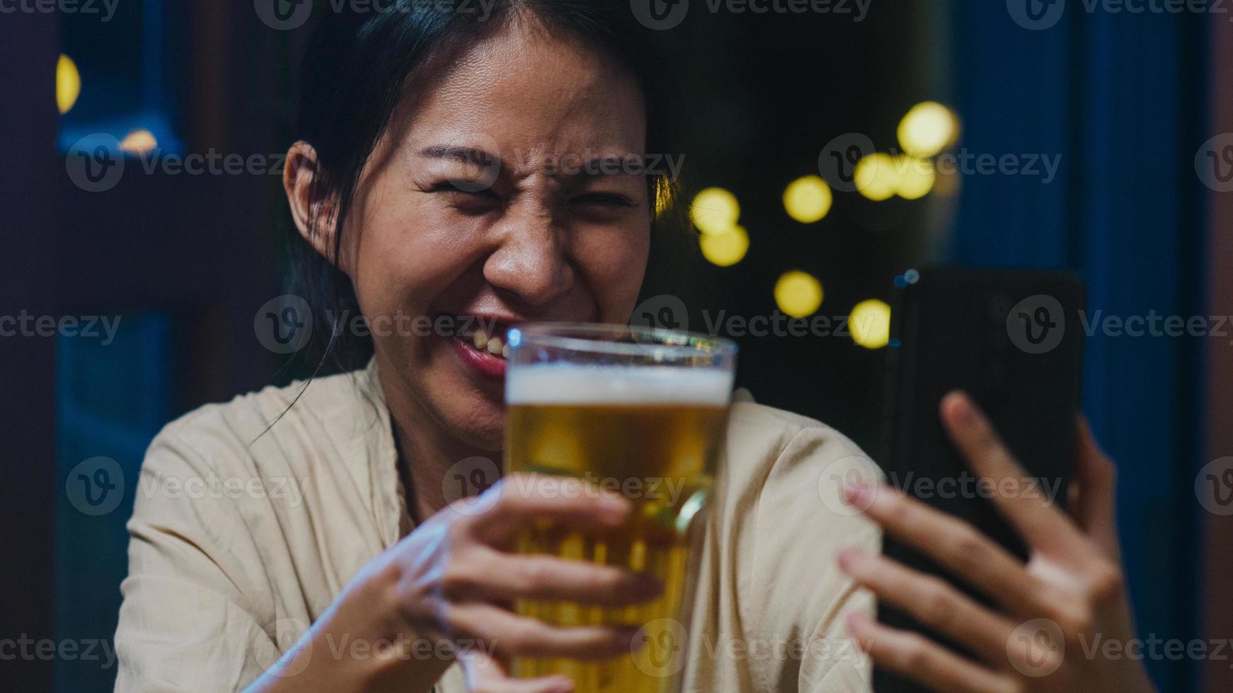 joven asiática bebiendo cerveza divirtiéndose feliz momento fiesta nocturna evento de año nuevo celebración en línea a través de videollamada por teléfono en casa por la noche. distanciamiento social, cuarentena para la prevención del coronavirus. foto