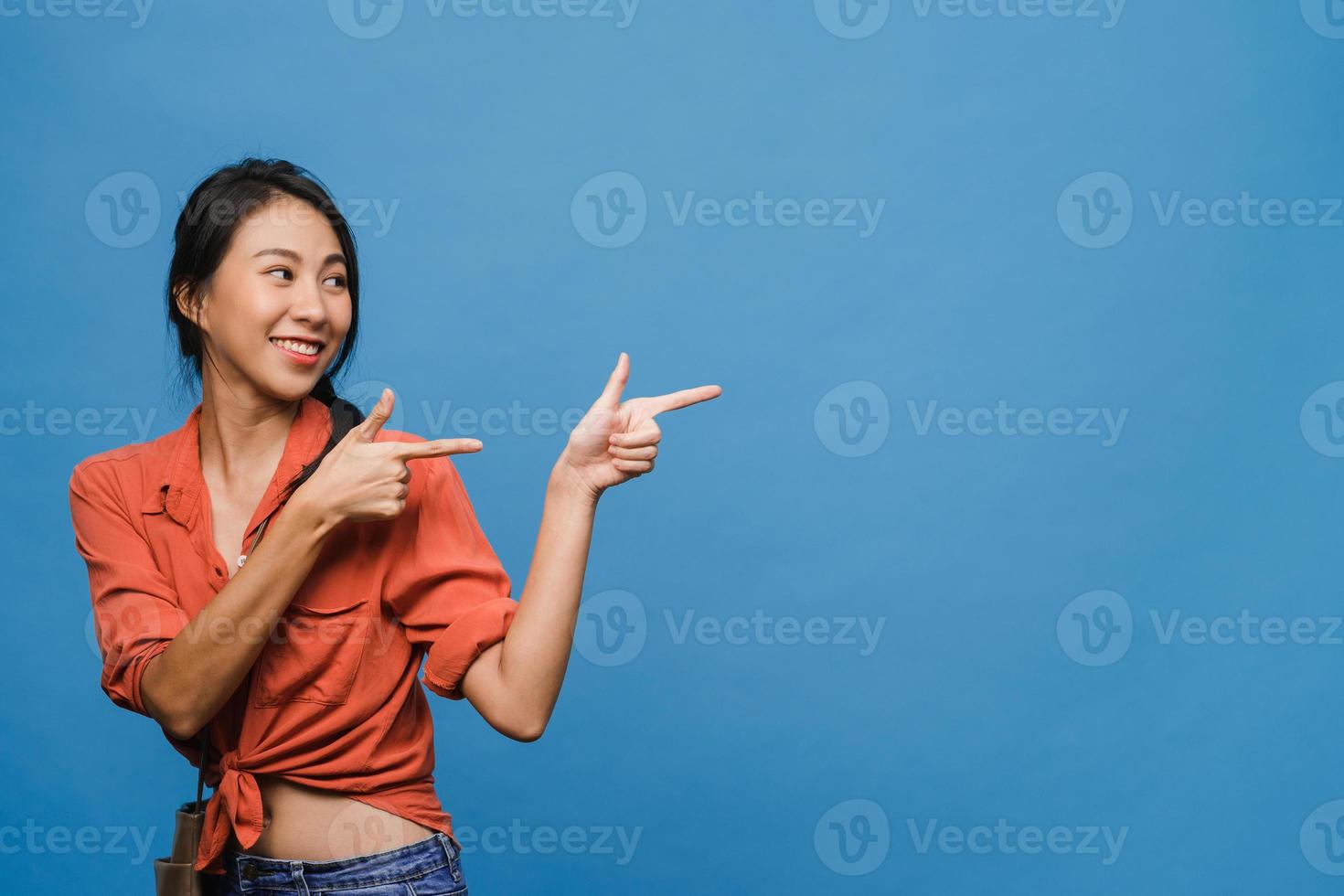 Portrait of young Asian lady smiling with cheerful expression, shows something amazing at blank space in casual clothing and standing isolated over blue background. Facial expression concept. photo