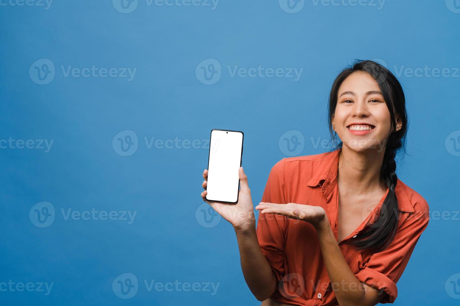 Young Asia lady show empty smartphone screen with positive expression, smiles broadly, dressed in casual clothing feeling happiness on blue background. Mobile phone with white screen in female hand. photo