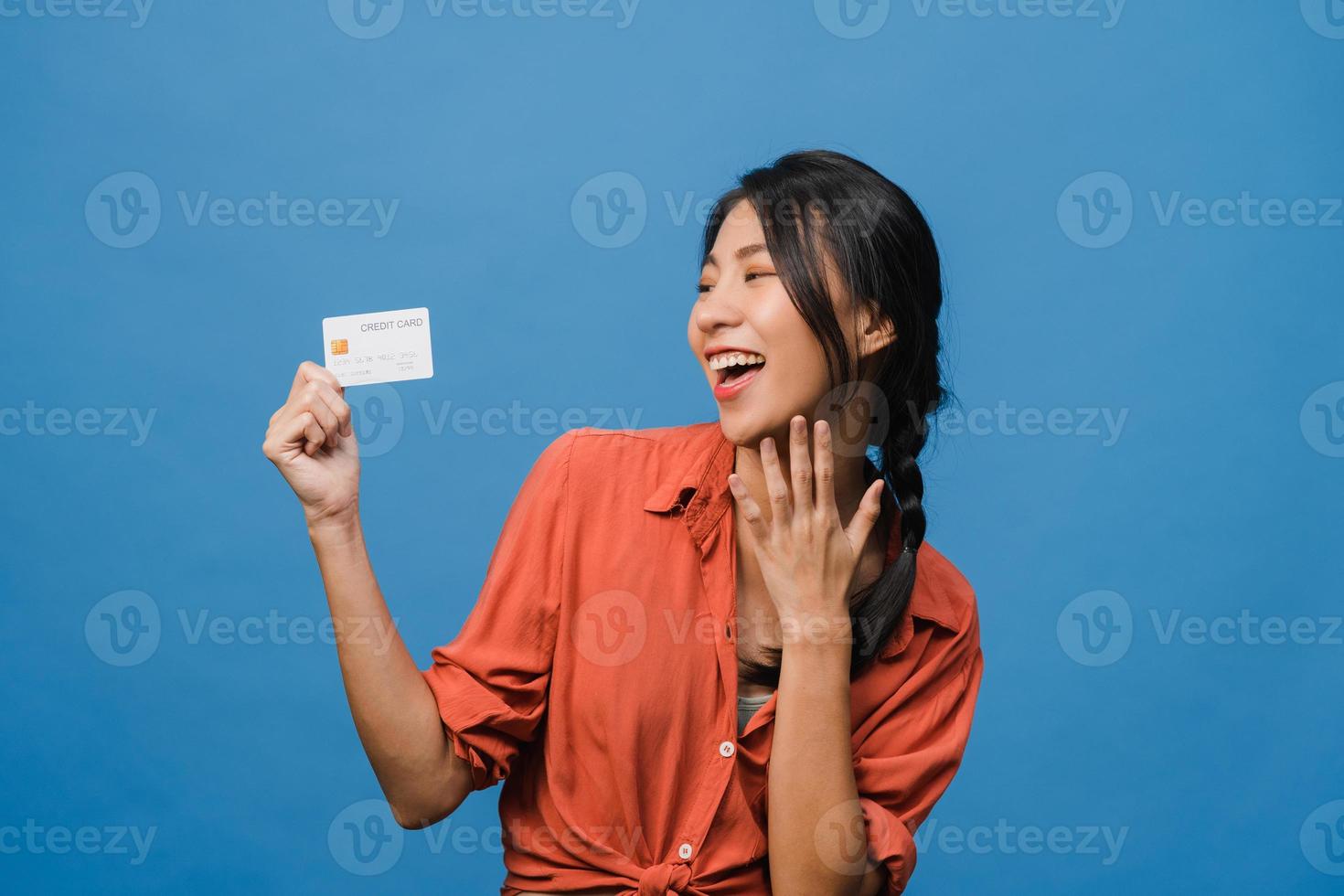 Joven mujer asiática muestra una tarjeta bancaria de crédito con expresión positiva, sonríe ampliamente, vestida con ropa informal sintiendo felicidad y parada aislada sobre fondo azul. concepto de expresión facial. foto