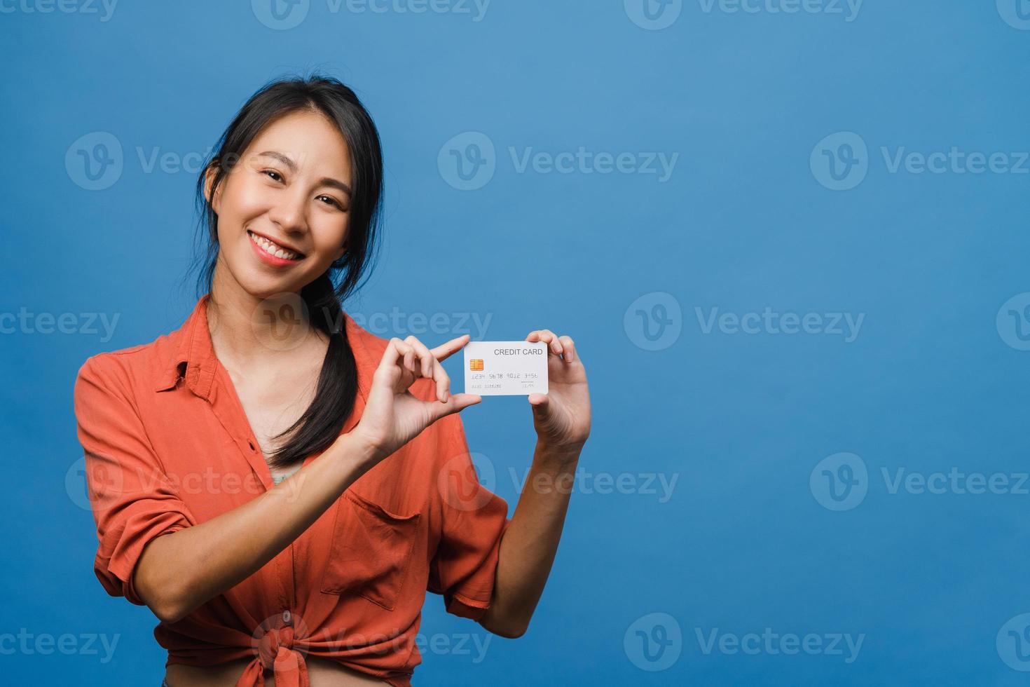 Joven mujer asiática muestra una tarjeta bancaria de crédito con expresión positiva, sonríe ampliamente, vestida con ropa informal sintiendo felicidad y parada aislada sobre fondo azul. concepto de expresión facial. foto