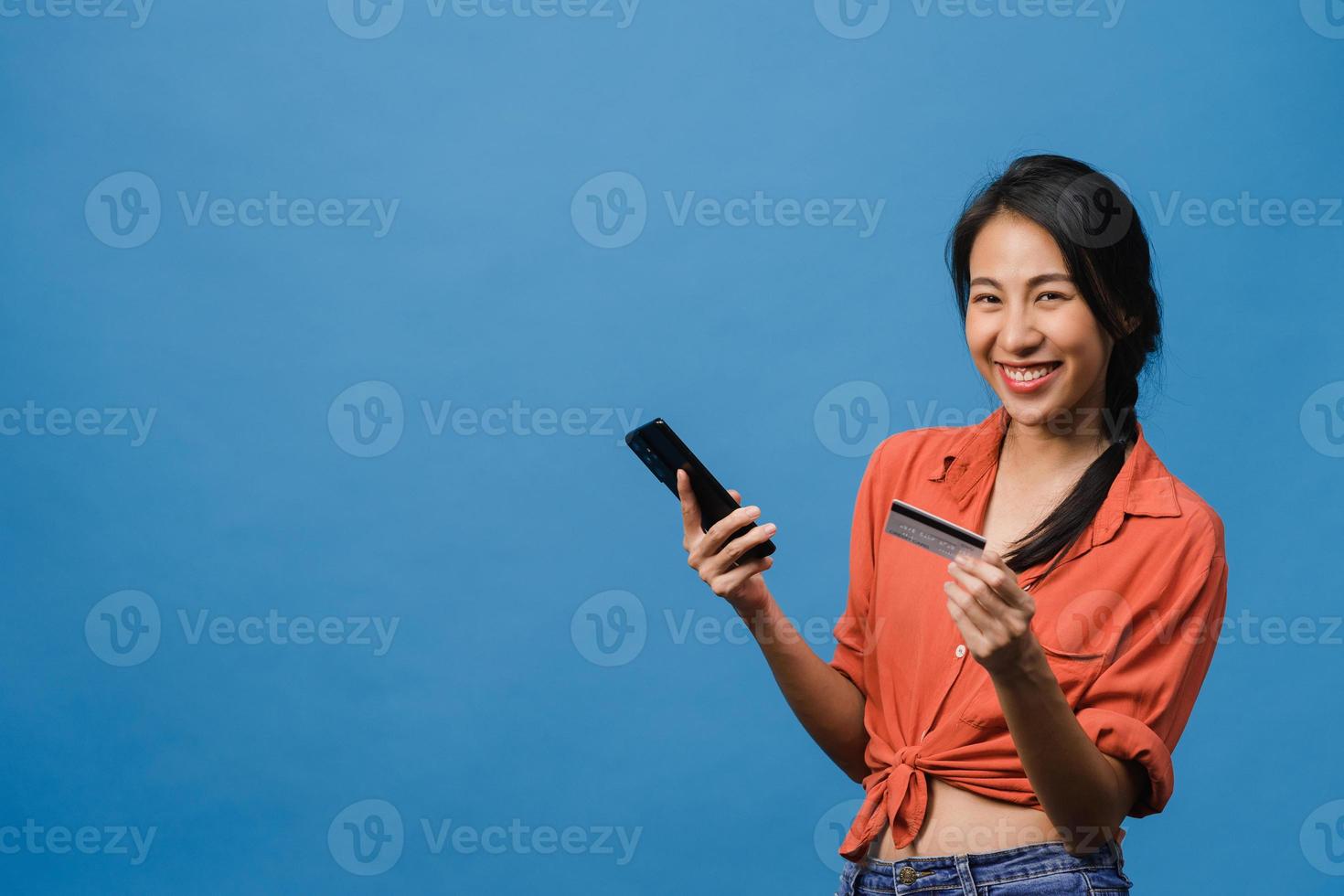 Young Asia lady using phone and credit bank card with positive expression, smiles broadly, dressed in casual clothing and stand isolated on blue background. Happy adorable glad woman rejoices success. photo