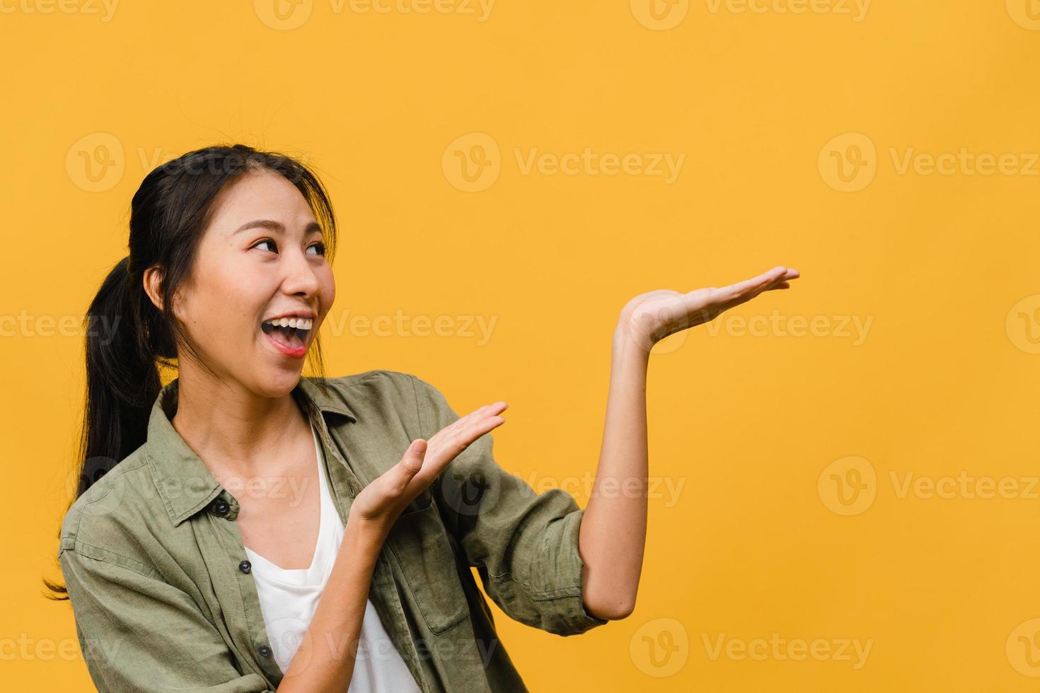 Retrato de joven asiática sonriendo con expresión alegre, muestra algo sorprendente en el espacio en blanco en ropa casual y de pie aislado sobre fondo amarillo. concepto de expresión facial. foto