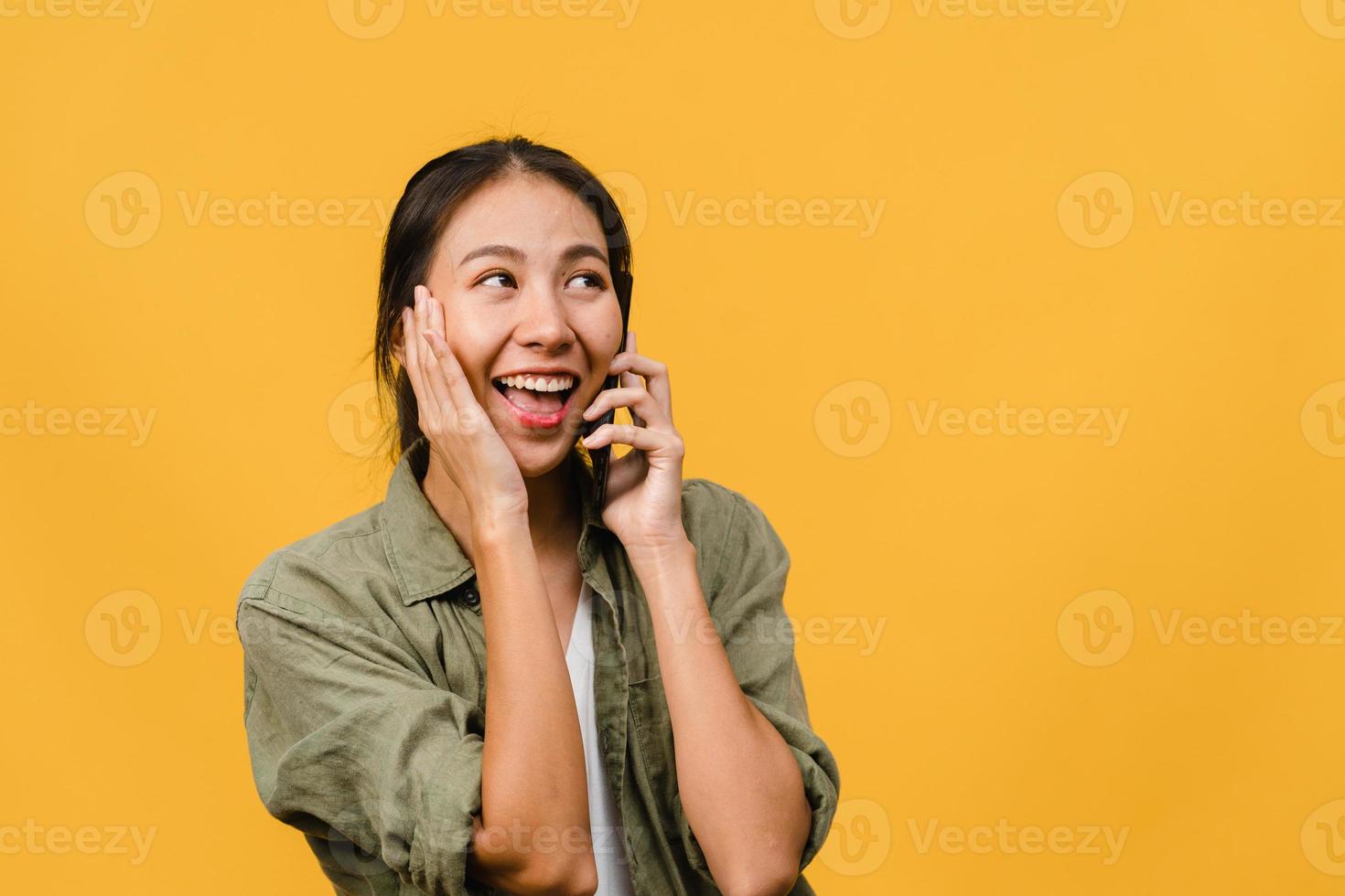 Young Asia lady talk by phone with positive expression, smile broadly, dressed in casual clothing feeling happiness and stand isolated on yellow background. Happy adorable glad woman rejoices success. photo