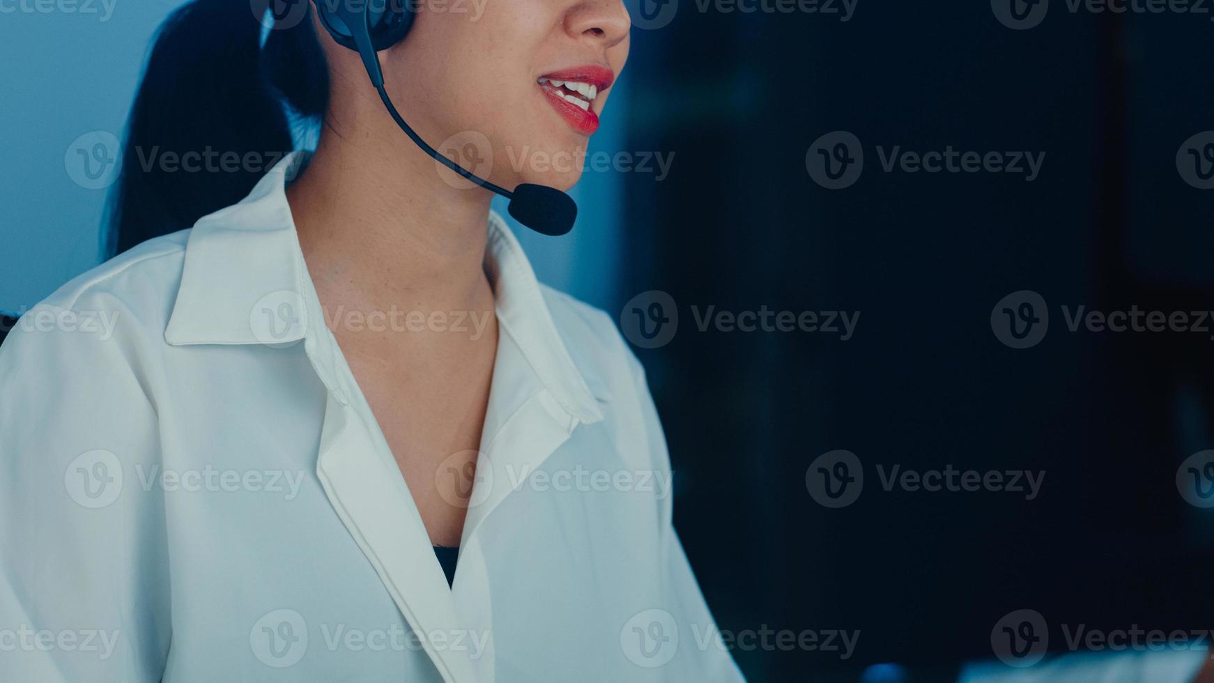 Millennial Asia young call center agent or customer support service executive using computer and microphone headset working technical support in late night office. Telemarketing or sales job concept. photo