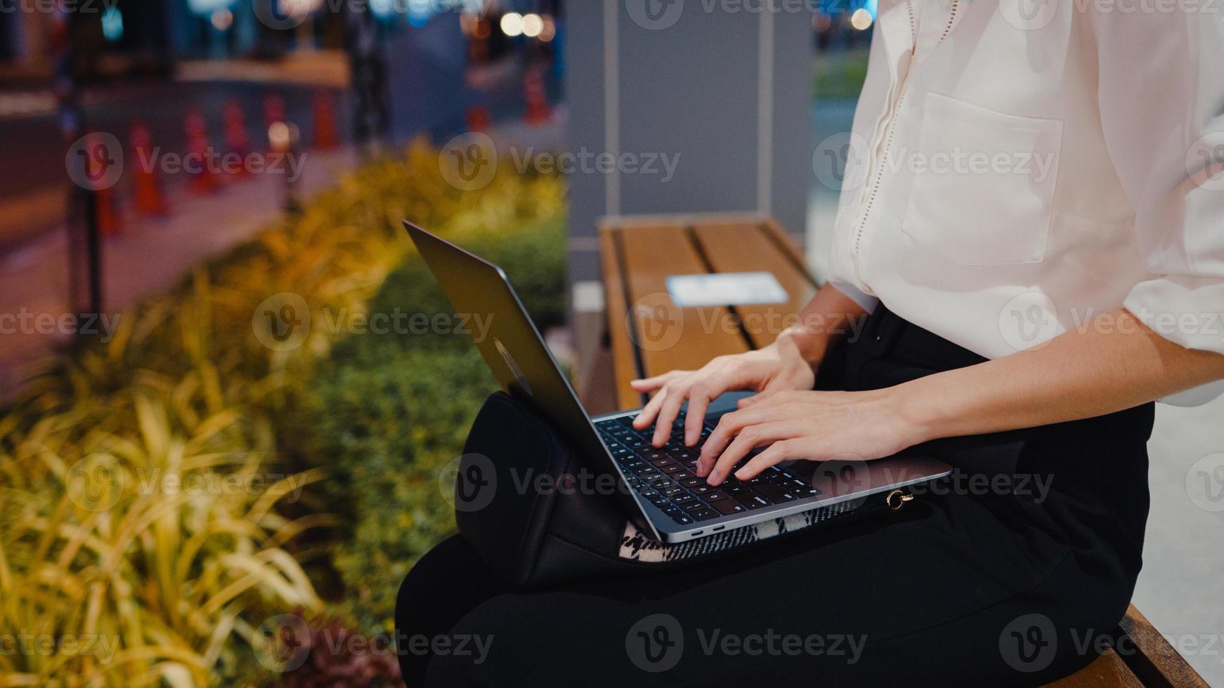 Exitosa empresaria asiática joven en ropa de oficina de moda usando laptop trabaja de forma remota mientras está sentada sola al aire libre en la ciudad moderna urbana en la noche. concepto de negocio en movimiento. foto