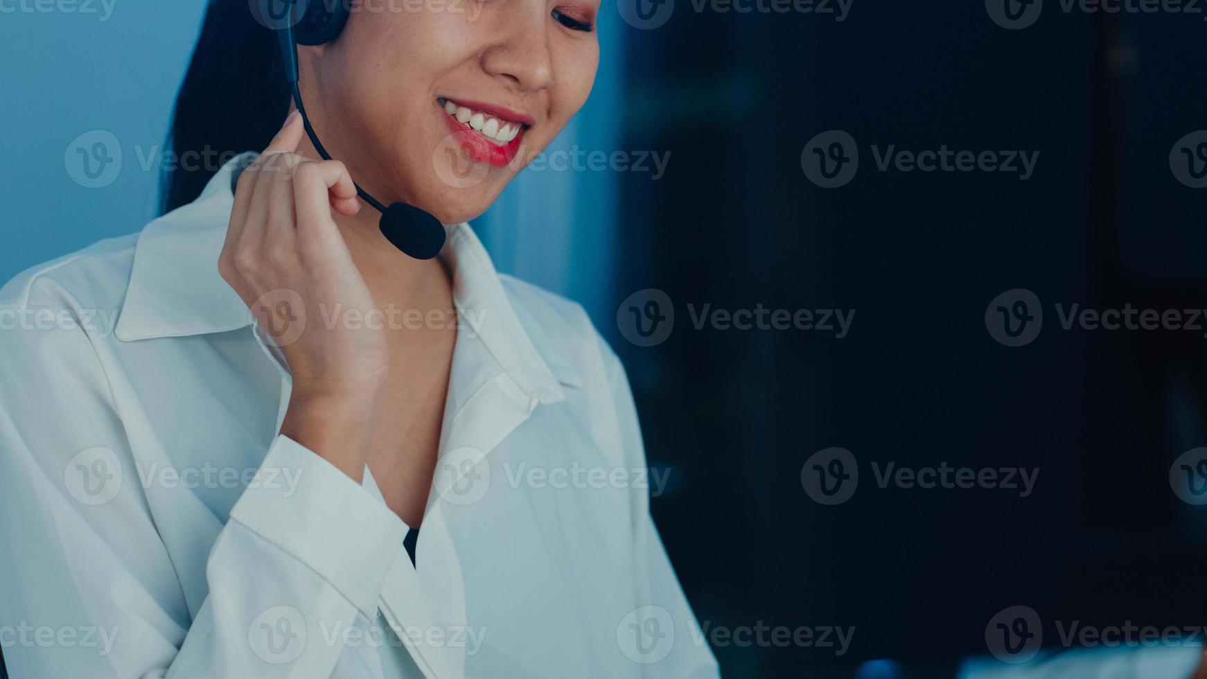 Millennial Asia young call center agent or customer support service executive using computer and microphone headset working technical support in late night office. Telemarketing or sales job concept. photo