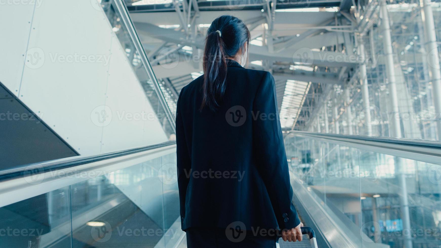 Asian business girl wear face mask drag luggage stand on escalator look around walk to terminal at international airport. Business commuter covid pandemic, Business travel social distancing concept. photo