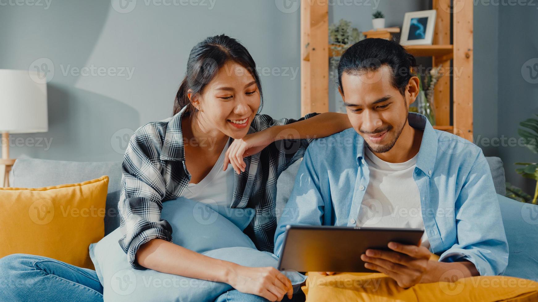 Feliz pareja joven y atractiva asiática, hombre y mujer, sentarse en el sofá, usar la tableta, comprar muebles en línea, decorar el hogar en la sala de estar de la nueva casa. joven casado mudanza concepto online comprador de casa. foto