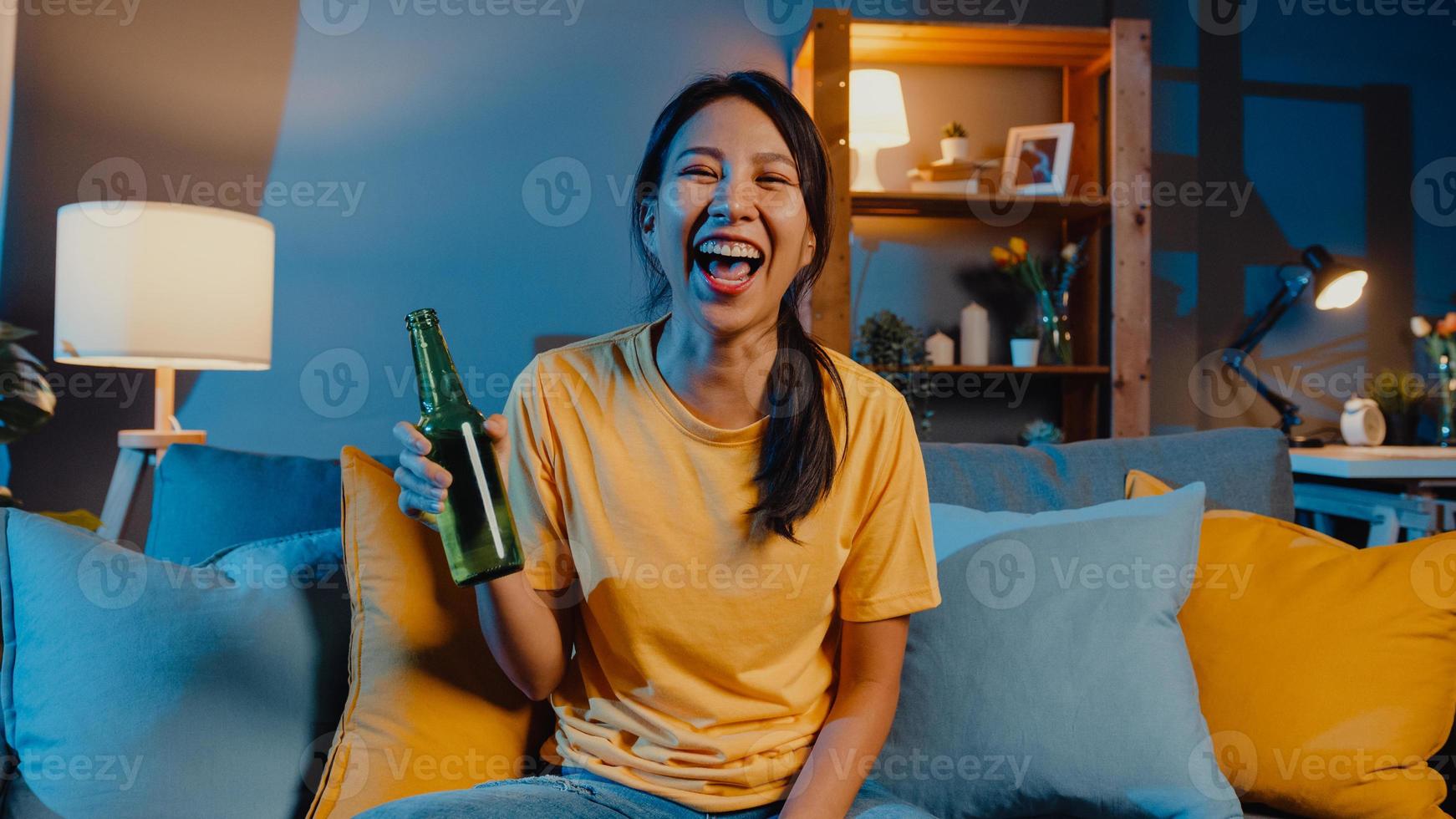 Happy young asian woman looking at camera enjoy night party event online with friends toast drink beer via video call online in living room at home, Stay at home quarantine, Social distancing concept. photo