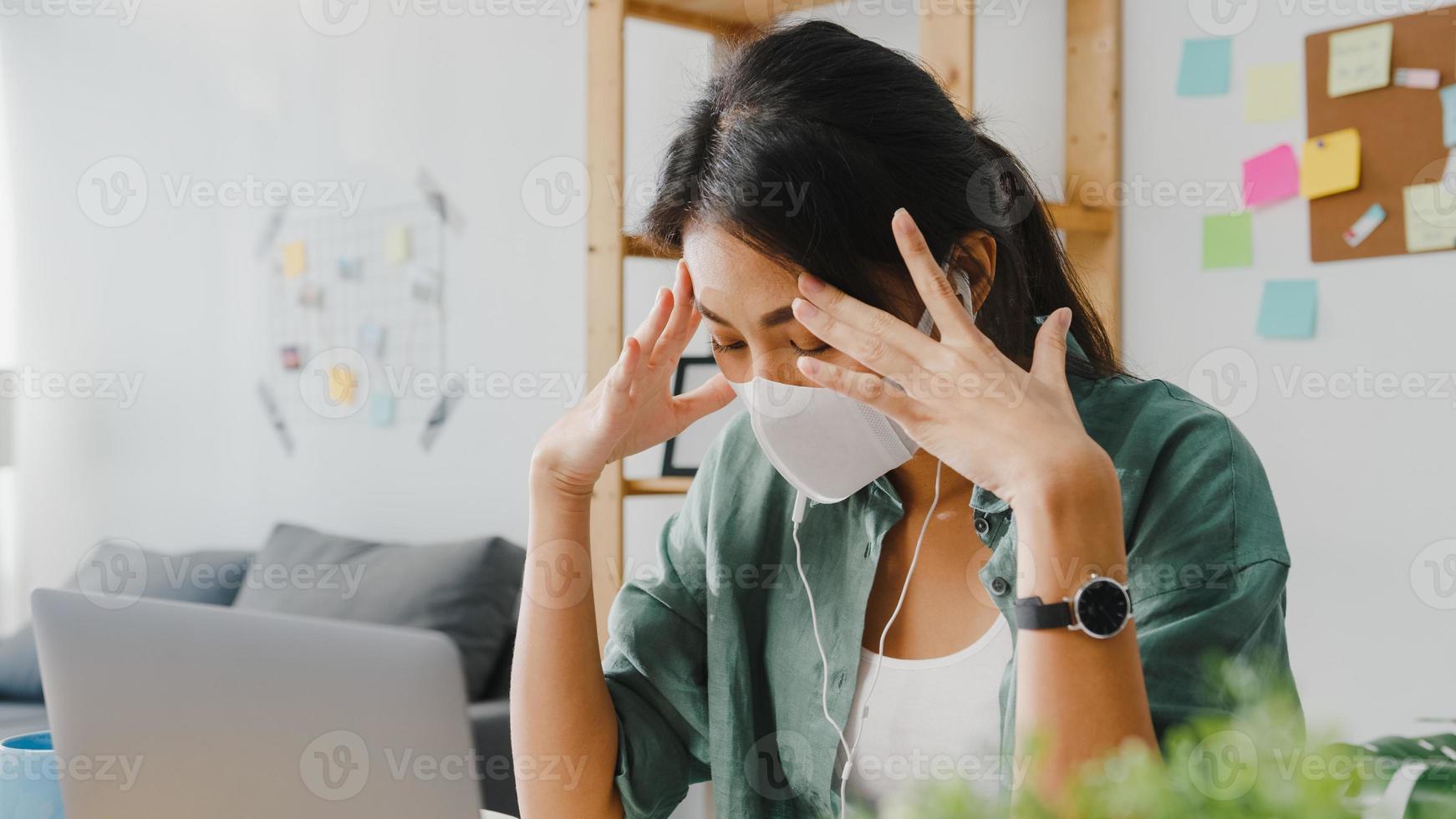 Asia businesswoman wearing medical face mask using laptop talk to colleagues about plan in video call while working from home at living room. Social distancing, quarantine for corona virus prevention. photo