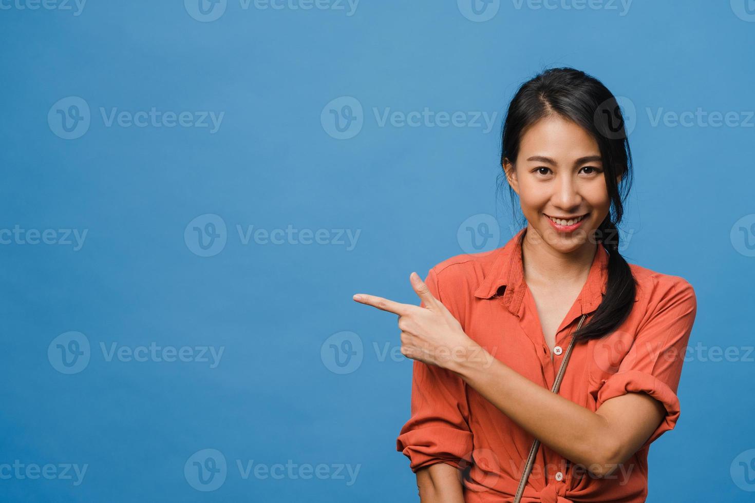 Portrait of young Asian lady smiling with cheerful expression, shows something amazing at blank space in casual cloth and looking at camera isolated over blue background. Facial expression concept. photo