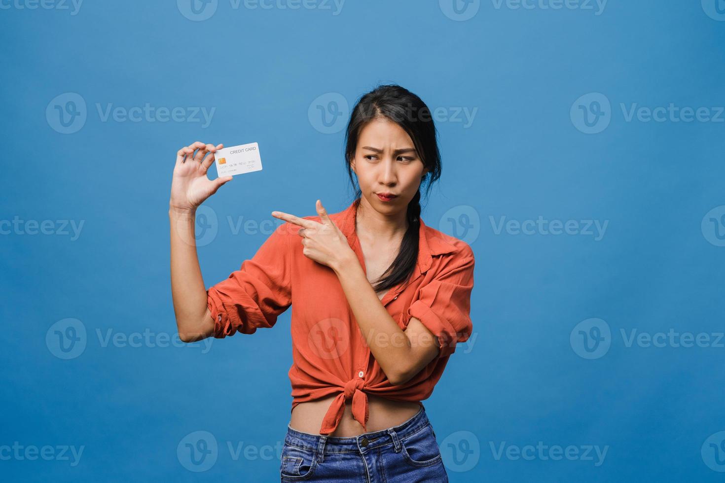 Joven mujer asiática muestra una tarjeta bancaria de crédito con expresión positiva, sonríe ampliamente, vestida con ropa informal sintiendo felicidad y parada aislada sobre fondo azul. concepto de expresión facial. foto