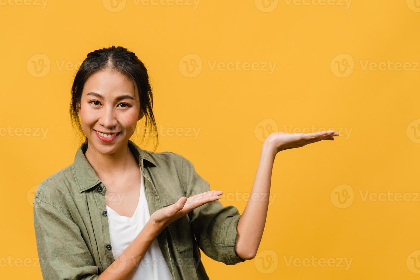 Portrait of young Asian lady smiling with cheerful expression, shows something amazing at blank space in casual cloth and looking at camera isolated over yellow background. Facial expression concept. photo
