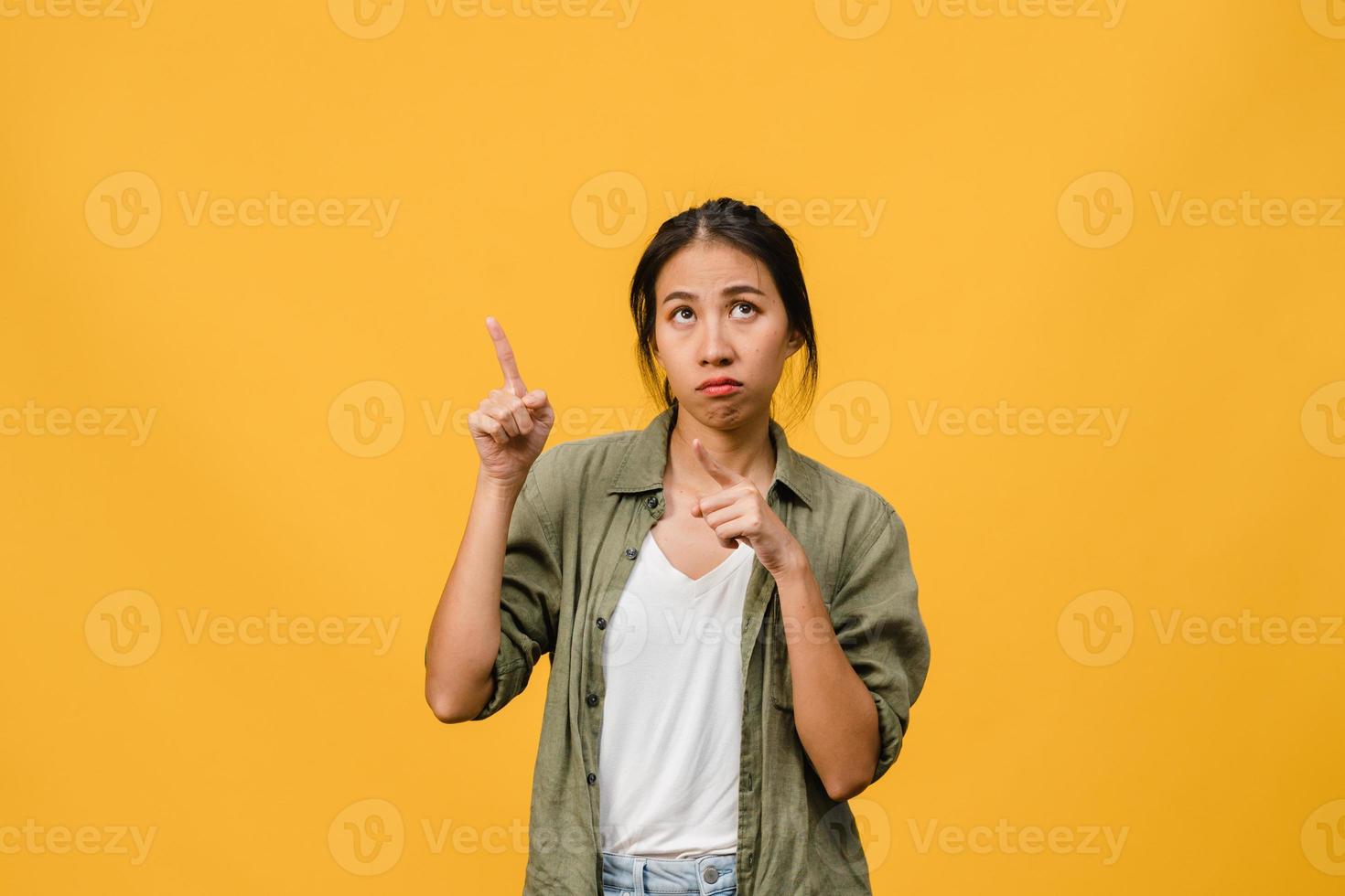 Joven mujer asiática muestra algo sorprendente en el espacio en blanco con expresión negativa, gritos emocionados, llorando enojado emocional en ropa casual aislada sobre fondo amarillo. concepto de expresión facial. foto