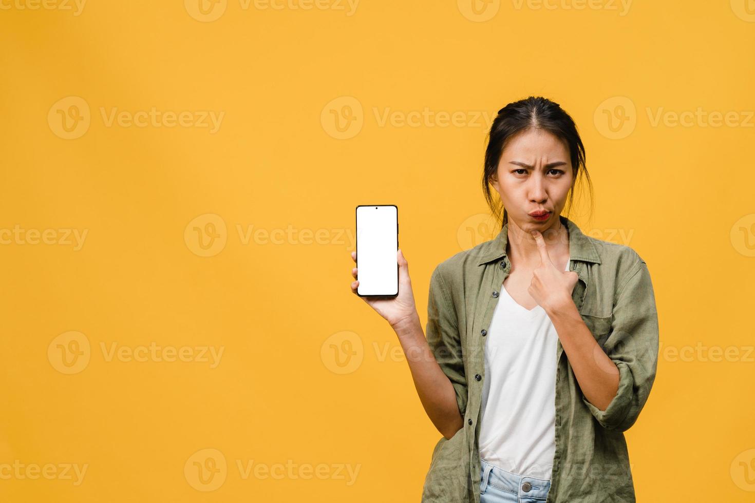 Joven asiática muestra la pantalla vacía del teléfono inteligente con expresión positiva, sonríe ampliamente, vestida con ropa casual sintiendo felicidad sobre fondo amarillo. teléfono móvil con pantalla en blanco en mano femenina. foto