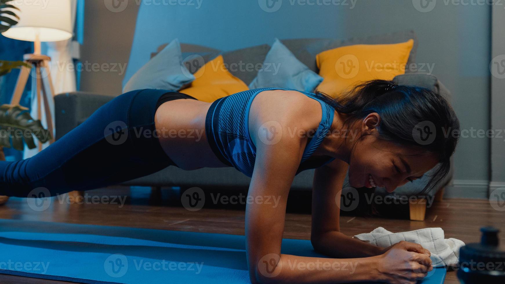 Jovencita asiática en ejercicios de ropa deportiva haciendo ejercicio y usando el teléfono para ver el video tutorial de yoga en la noche en casa. entrenamiento a distancia con entrenador personal, distancia social, concepto de educación en línea. foto