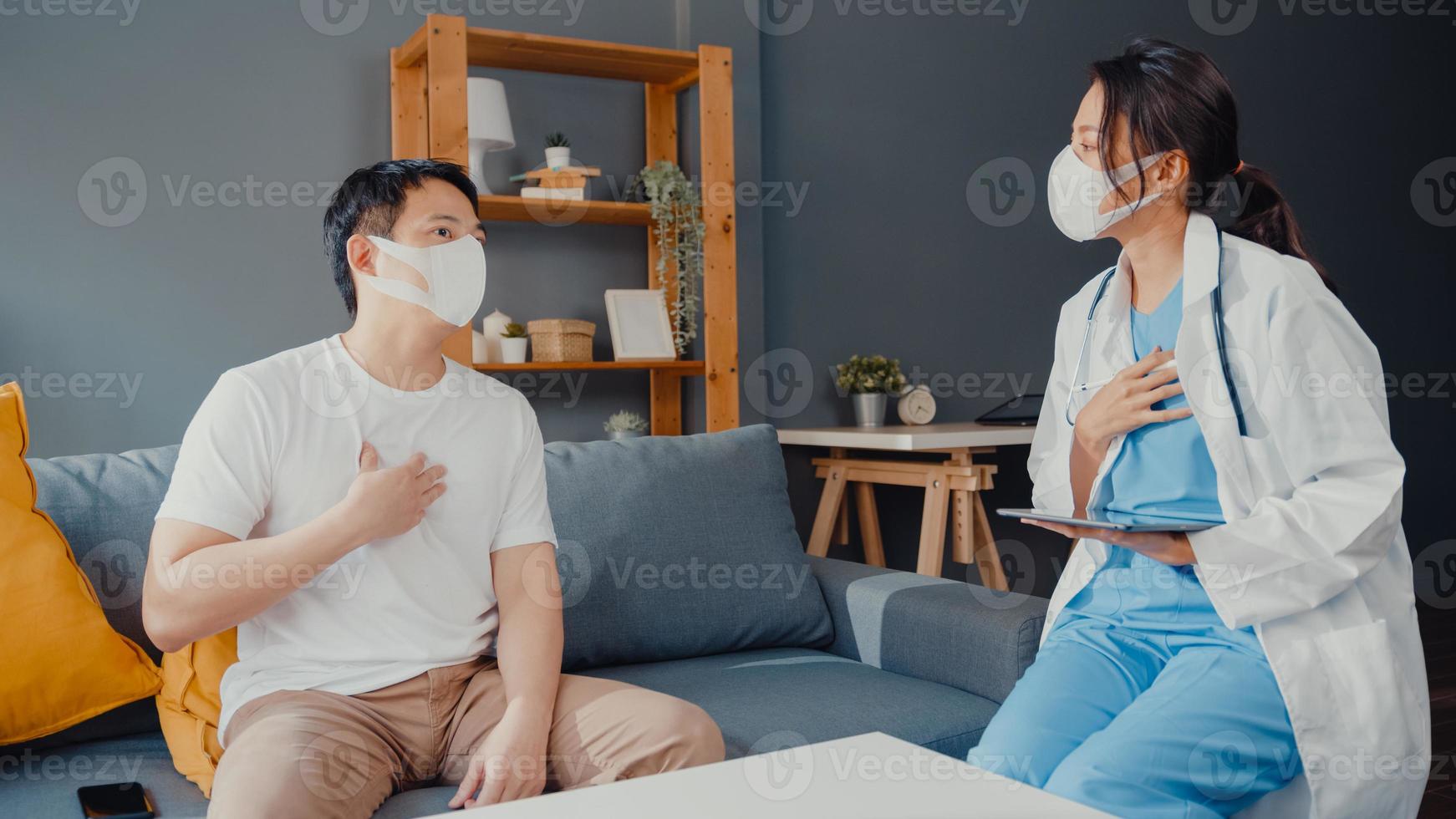 Young Asia female physician doctor wear face mask using digital tablet sharing good health test news with happy male patient sit on couch in house. Medical insurance, Visit patient at home concept. photo