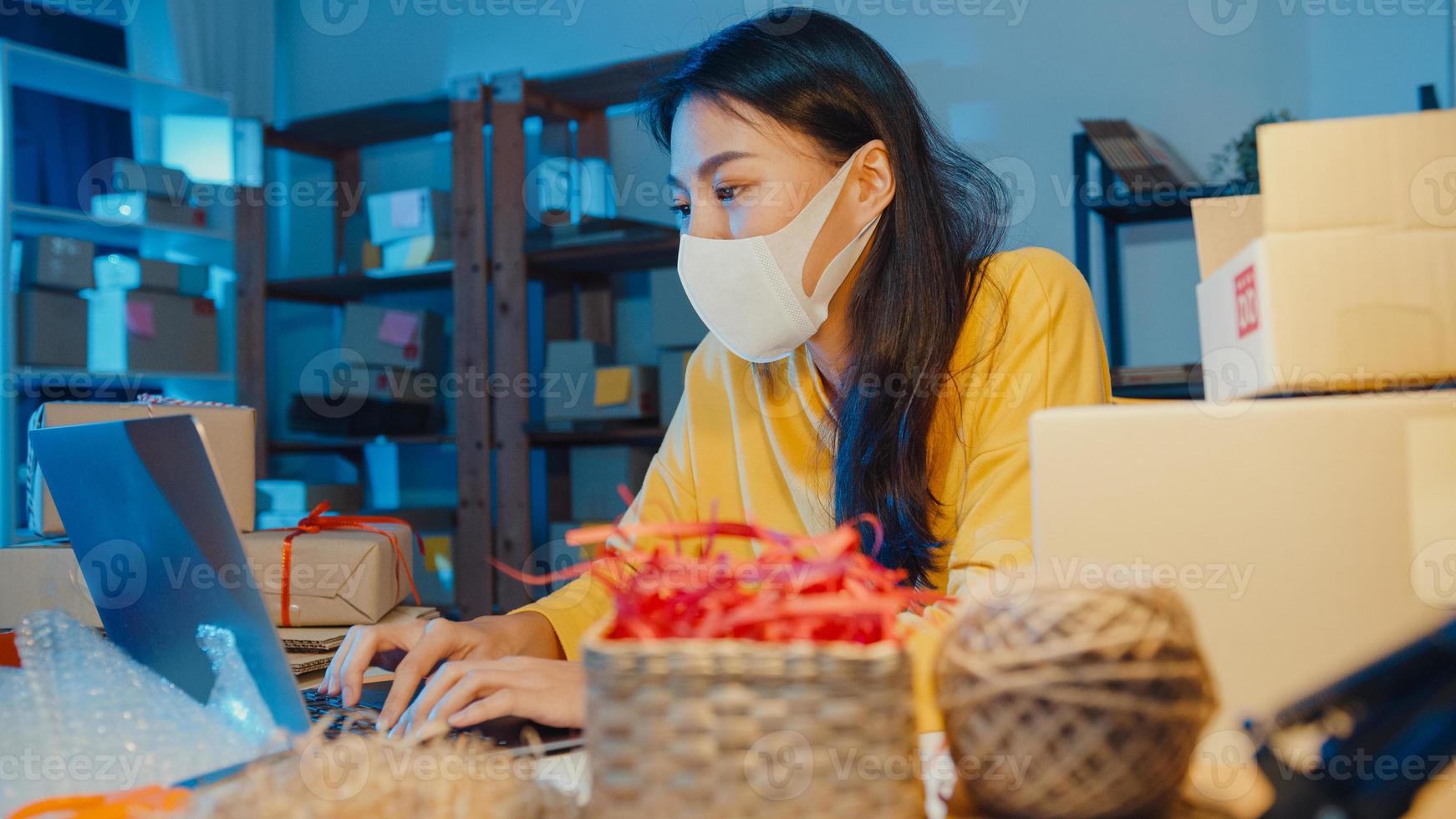 Young Asia businesswoman wear face mask check purchase order and reply client inbox chat on laptop in home office at night. Small business owner, online market delivery, lifestyle freelance concept. photo