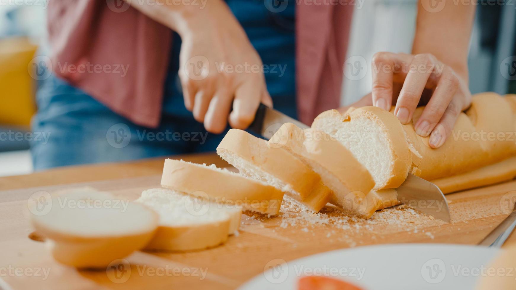 Manos de joven mujer asiática chef sosteniendo un cuchillo cortando pan integral sobre tabla de madera en la mesa de la cocina en casa. producción de pan casero fresco, alimentación saludable y concepto de panadería tradicional. foto