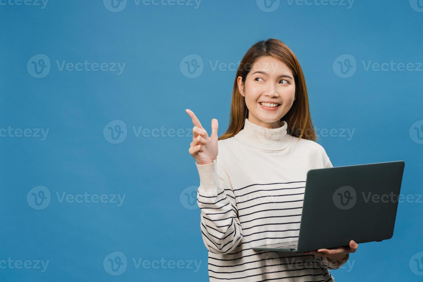 Jovencita asiática usando laptop con expresión positiva, sonríe ampliamente, vestida con ropa casual sintiendo felicidad y parada aislada sobre fondo azul. feliz adorable mujer alegre se regocija con el éxito. foto