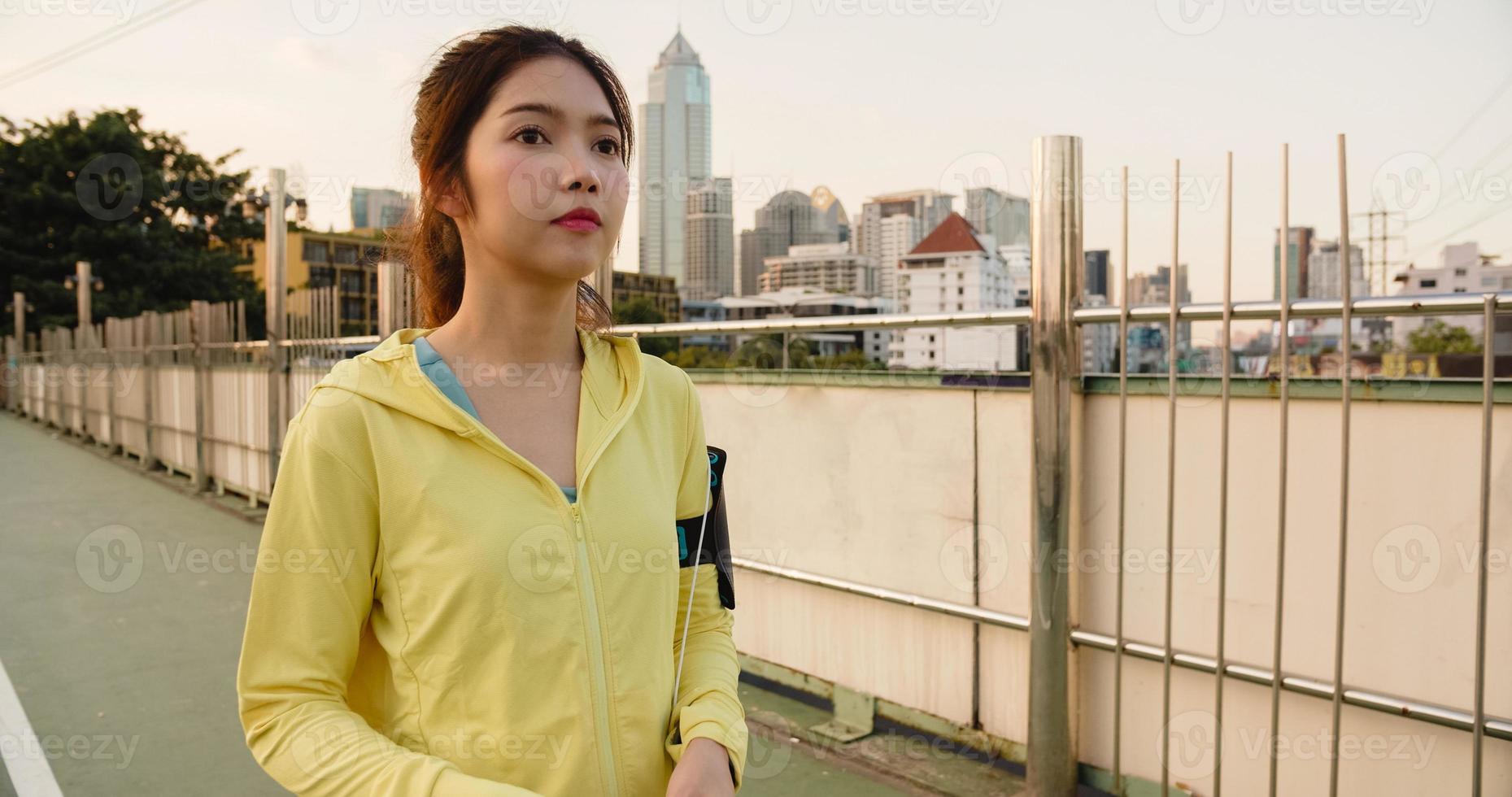 Beautiful young Asia athlete lady exercises using smartphone for listen to music while running in urban environment. Korean teen girl wearing sports clothes on walkway bridge in early morning. photo