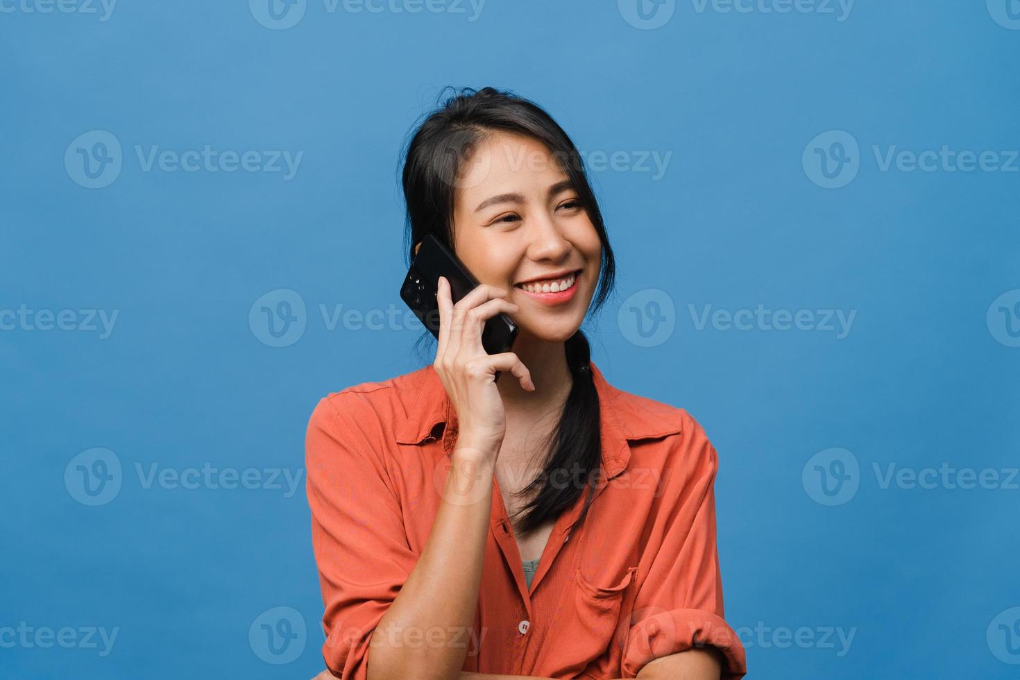Young Asia lady talk by phone with positive expression, smile broadly, dressed in casual clothing feeling happiness and stand isolated on blue background. Happy adorable glad woman rejoices success. photo