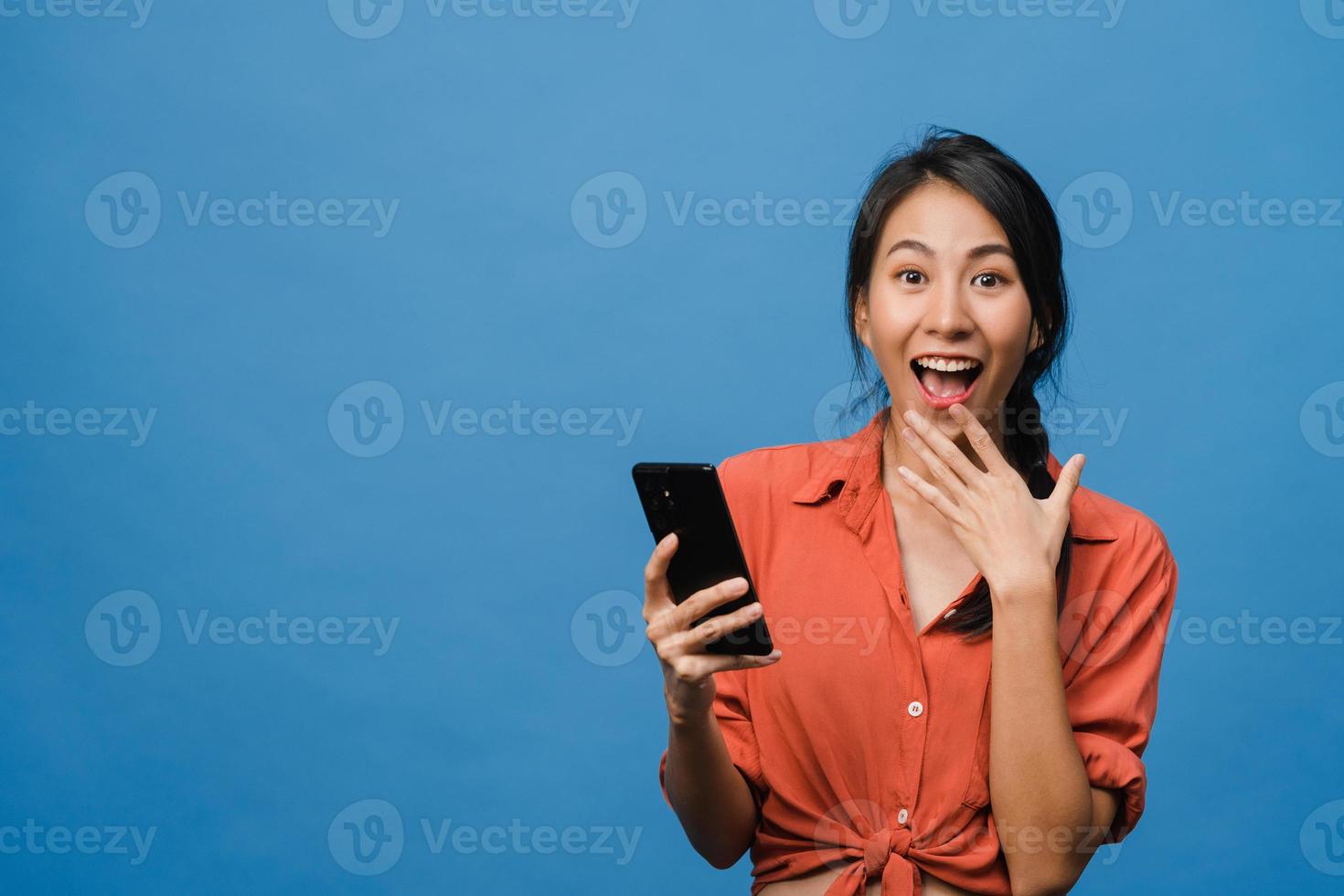 Sorprendió a la joven asiática que usa el teléfono móvil con expresión positiva, sonríe ampliamente, vestida con ropa informal y mirando a la cámara sobre fondo azul. feliz adorable mujer alegre se regocija con el éxito. foto