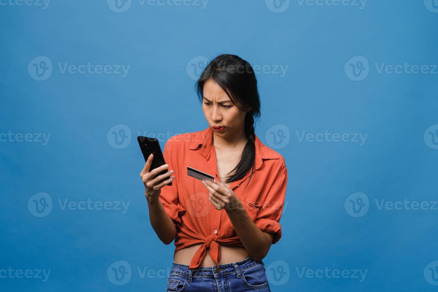 Jovencita asiática usando teléfono y tarjeta bancaria de crédito con expresión negativa, gritos emocionados, gritos emocionales enojados en ropa casual y de pie aislado sobre fondo azul. concepto de expresión facial. foto