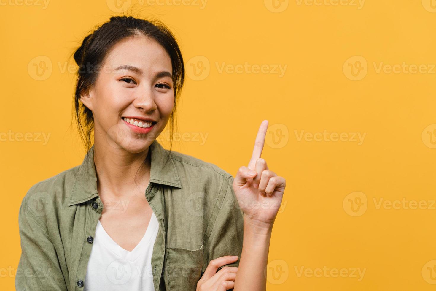 Portrait of young Asian lady smiling with cheerful expression, shows something amazing at blank space in casual cloth and looking at camera isolated over yellow background. Facial expression concept. photo