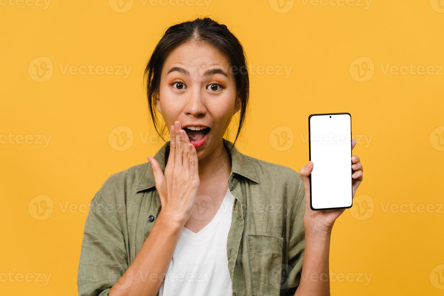 Joven asiática muestra la pantalla vacía del teléfono inteligente con expresión positiva, sonríe ampliamente, vestida con ropa casual sintiendo felicidad sobre fondo amarillo. teléfono móvil con pantalla en blanco en mano femenina. foto