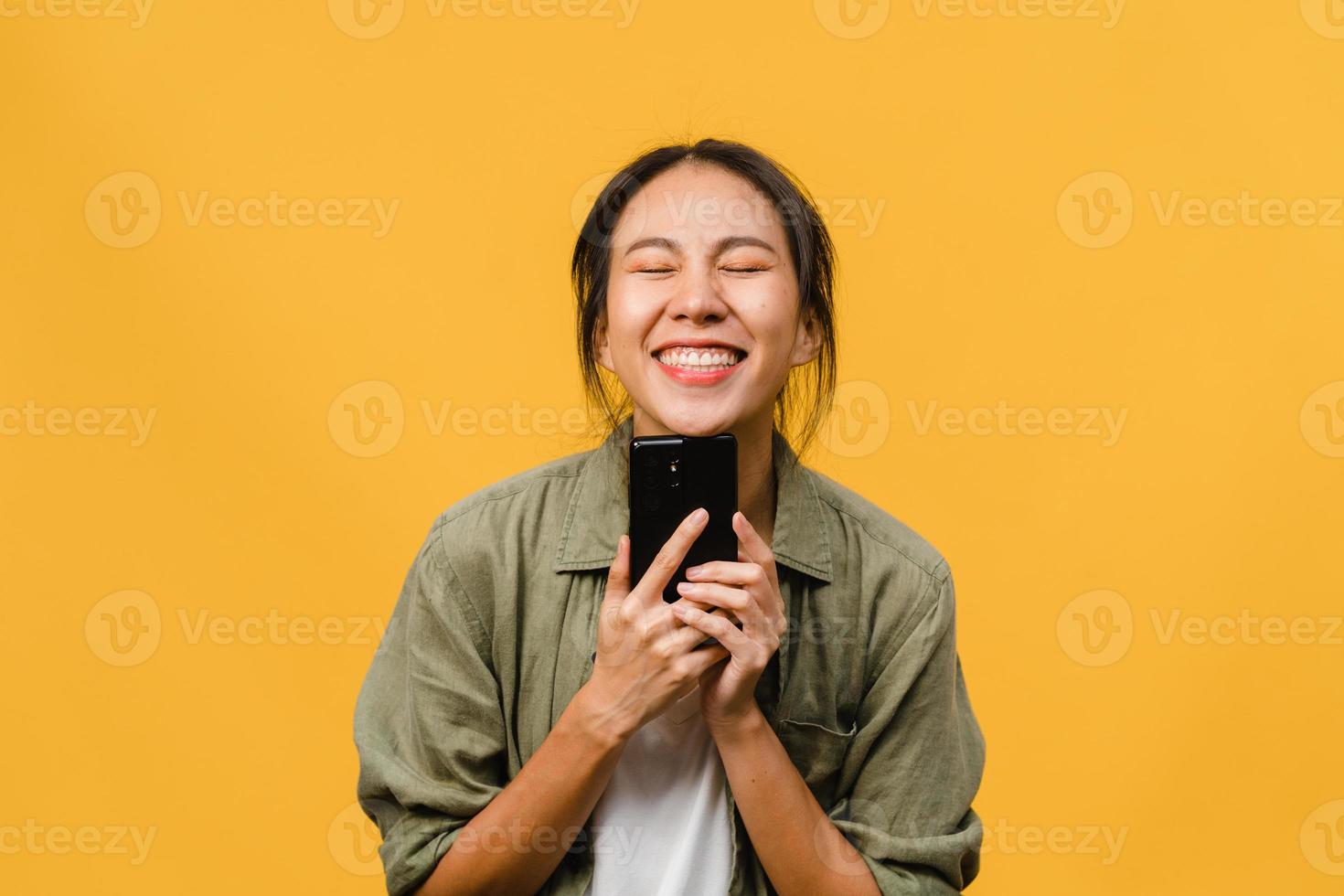 Jovencita asiática que usa el teléfono con expresión positiva, sonríe ampliamente, vestida con ropa casual sintiendo felicidad y parada aislada sobre fondo amarillo. feliz adorable mujer alegre se regocija con el éxito. foto