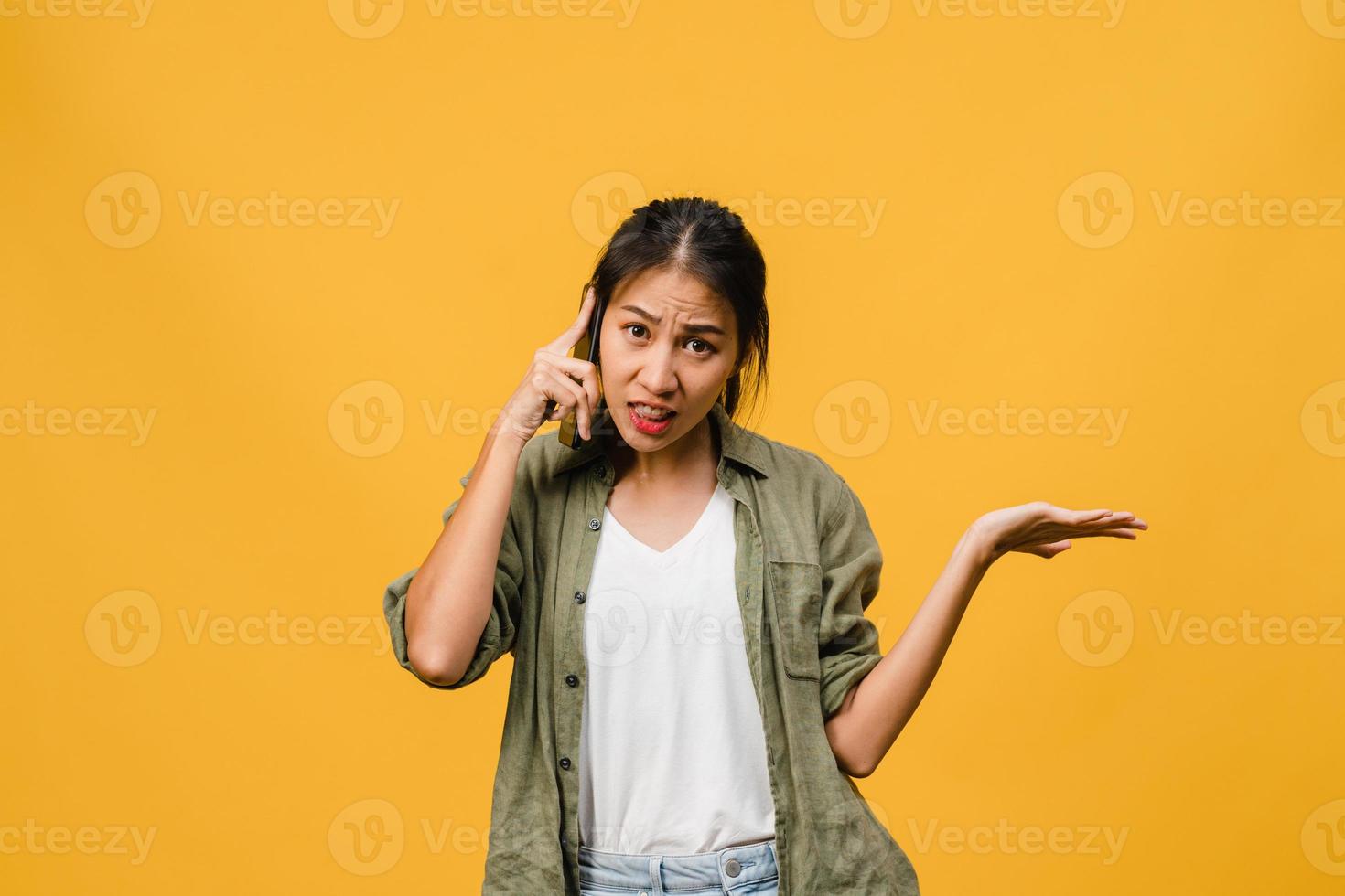 jovencita asiática habla por teléfono con expresión negativa, gritos emocionados, gritos emocionales enojados en ropa casual y se para aislado sobre fondo amarillo con espacio de copia en blanco. concepto de expresión facial. foto