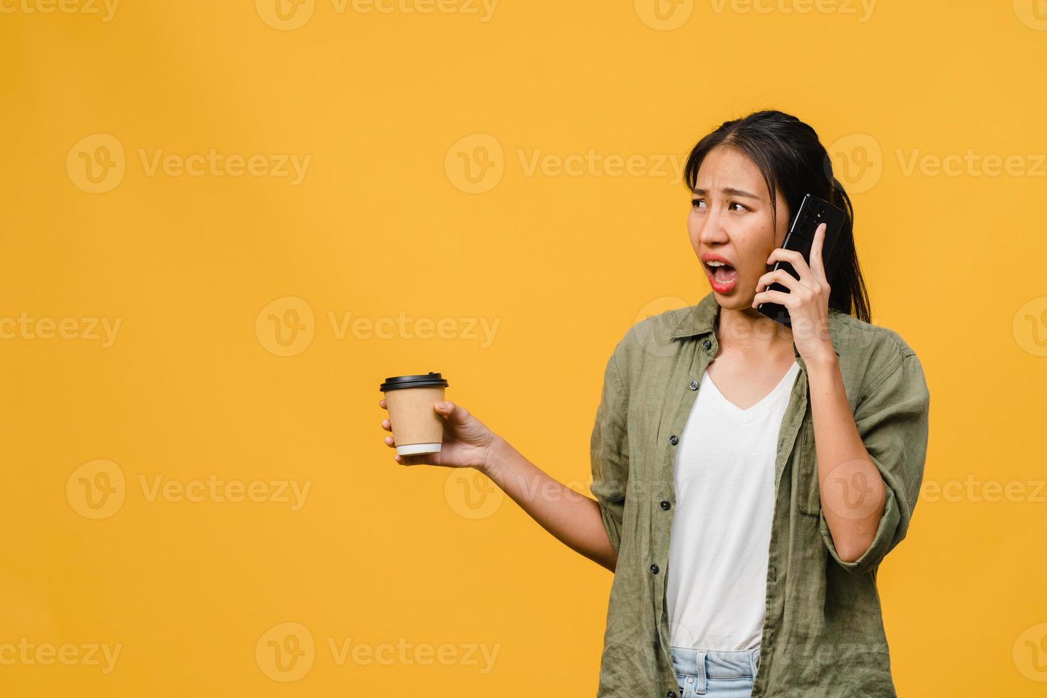 Jovencita asiática habla por teléfono y sostiene la taza de café con expresión negativa, gritos emocionados, llora enojado emocionalmente en ropa casual y se para aislado sobre fondo amarillo. concepto de expresión facial. foto