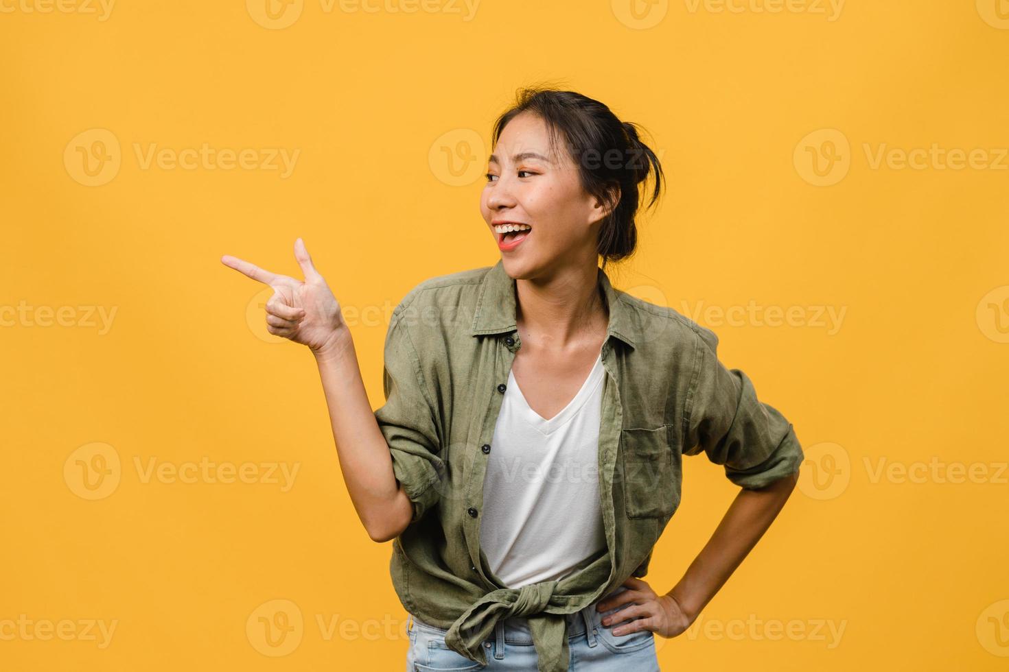 Retrato de joven asiática sonriendo con expresión alegre, muestra algo sorprendente en el espacio en blanco en ropa casual y de pie aislado sobre fondo amarillo. concepto de expresión facial. foto