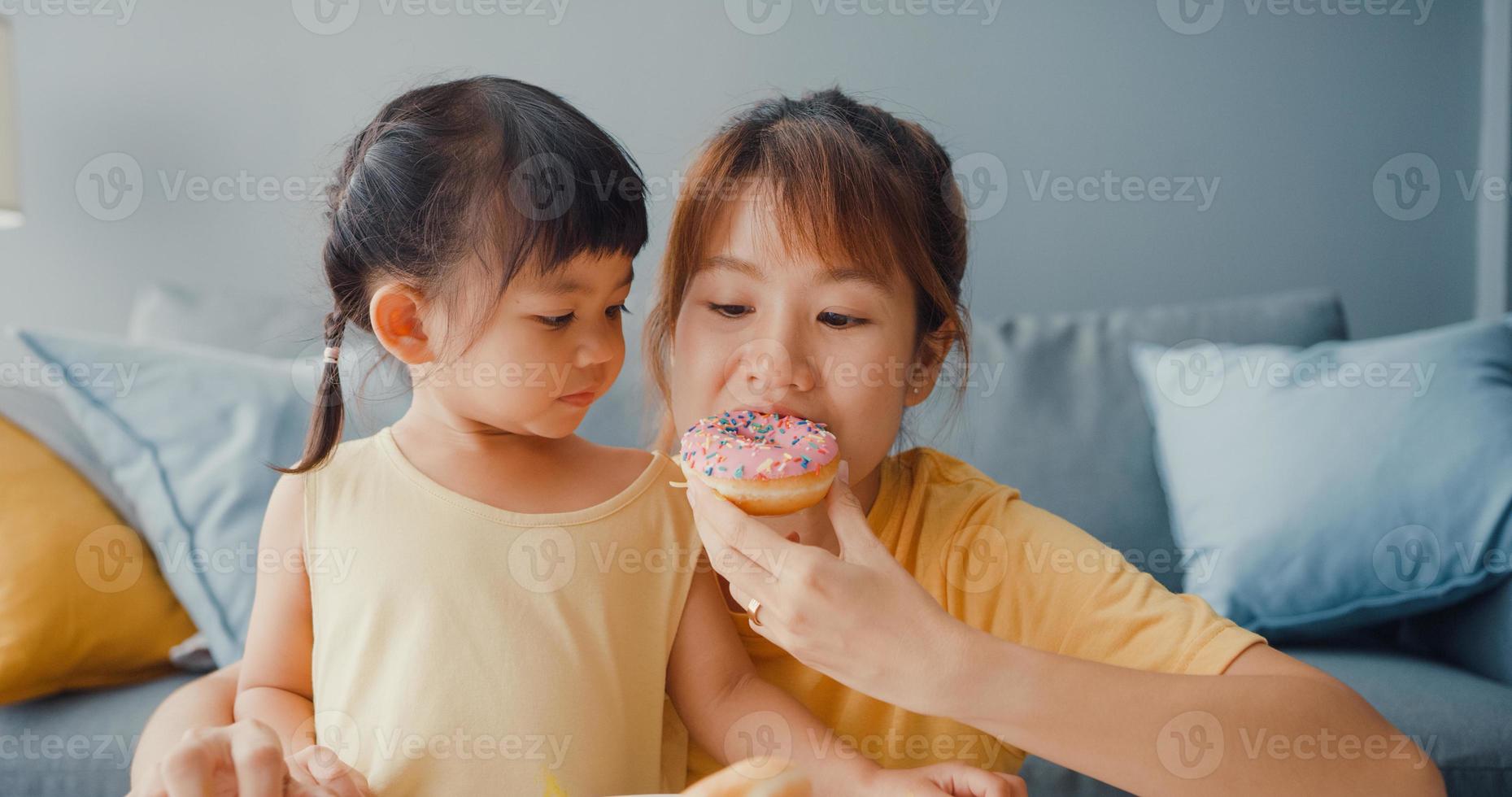 Happy cheerful Asia family mom and toddler girl eating donuts and having fun relax enjoy on couch in living room at house. Spending time together, Social distance, Quarantine for coronavirus. photo