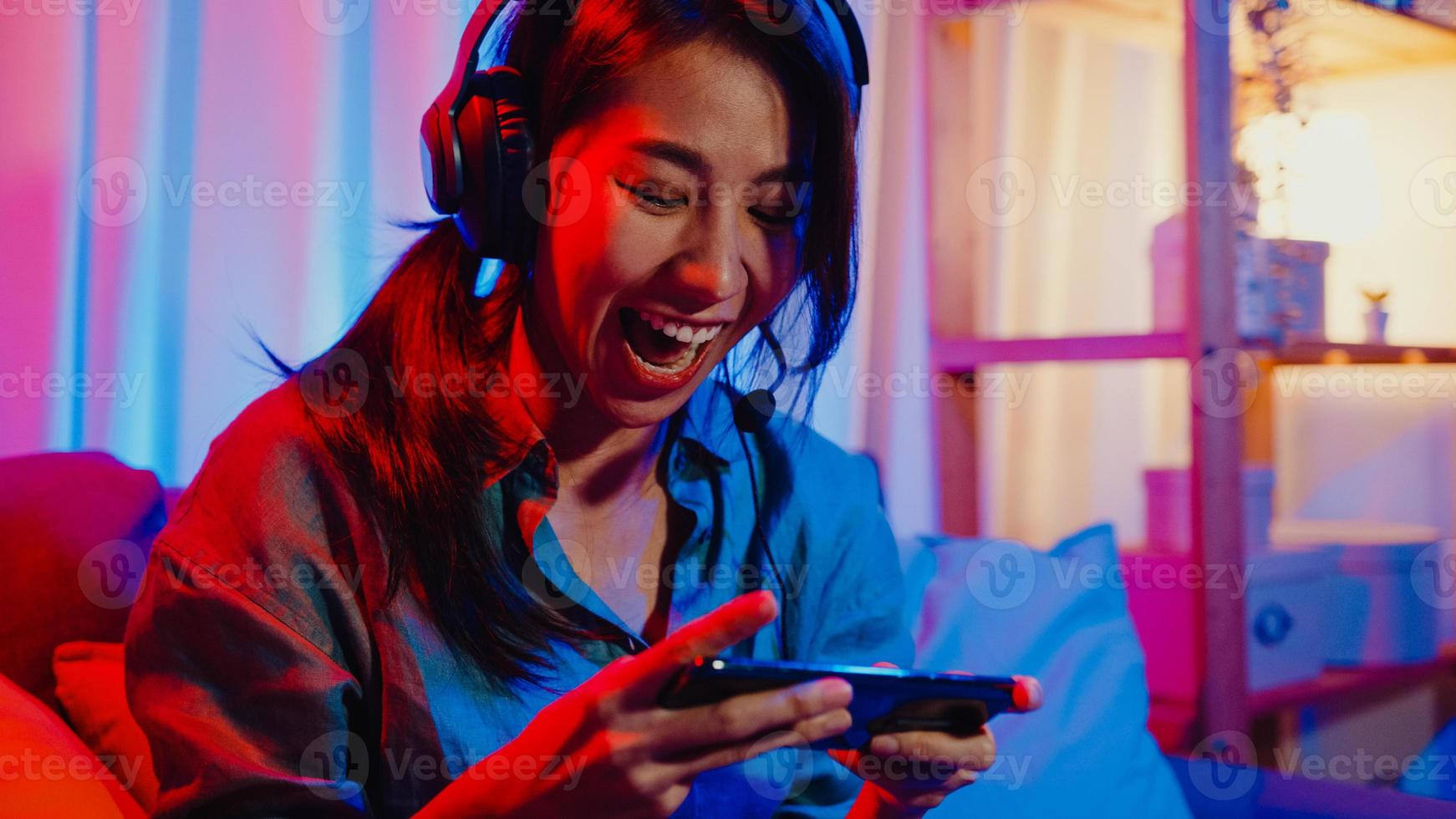 Happy asia girl gamer wear headphone competition video game online with smartphone excited talk with friend sit on couch in colorful neon lights living room at home, Home quarantine activity concept. photo