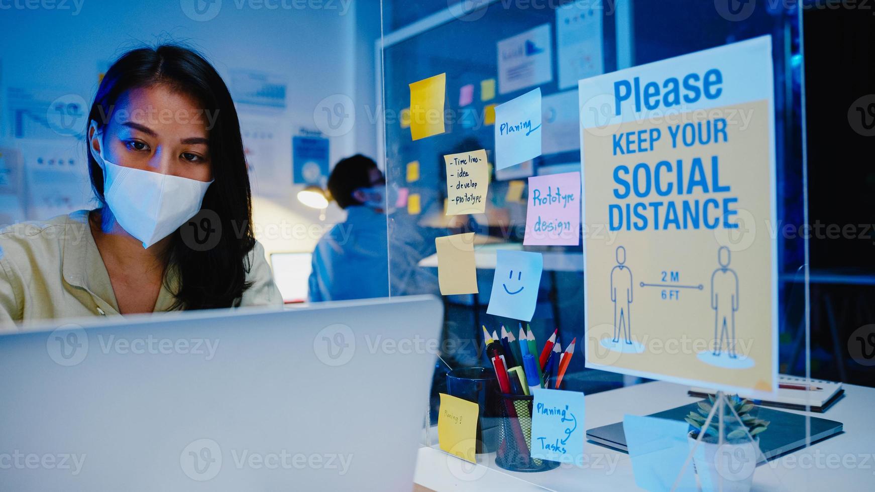 Feliz empresaria asiática con mascarilla médica para el distanciamiento social en una nueva situación normal para la prevención de virus mientras usa la computadora portátil en el trabajo en la noche de la oficina. vida y trabajo después del coronavirus. foto