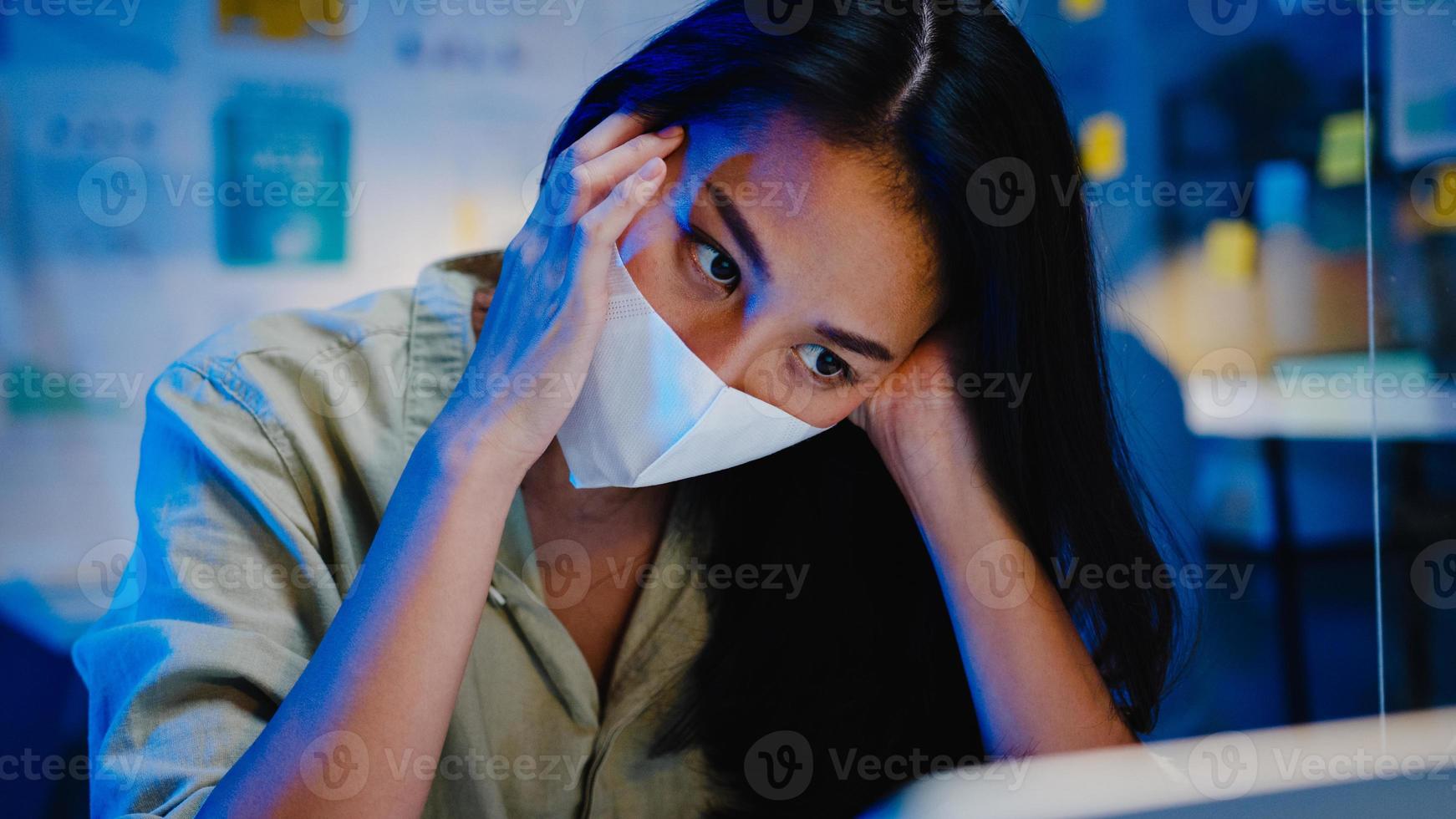 Freelance Asia women wear face mask using laptop hard work at new normal office. Working from home overload at night, self isolation, social distancing, quarantine for corona virus prevention. photo