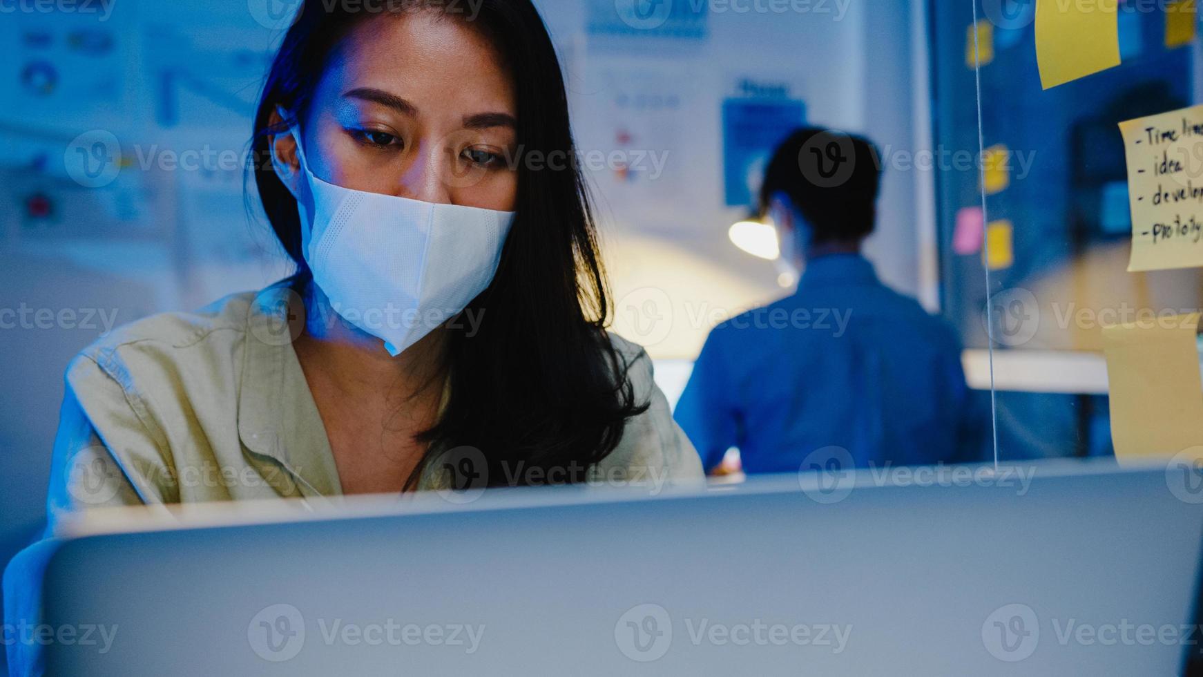 Feliz empresaria asiática con mascarilla médica para el distanciamiento social en una nueva situación normal para la prevención de virus mientras usa la computadora portátil en el trabajo en la noche de la oficina. vida y trabajo después del coronavirus. foto