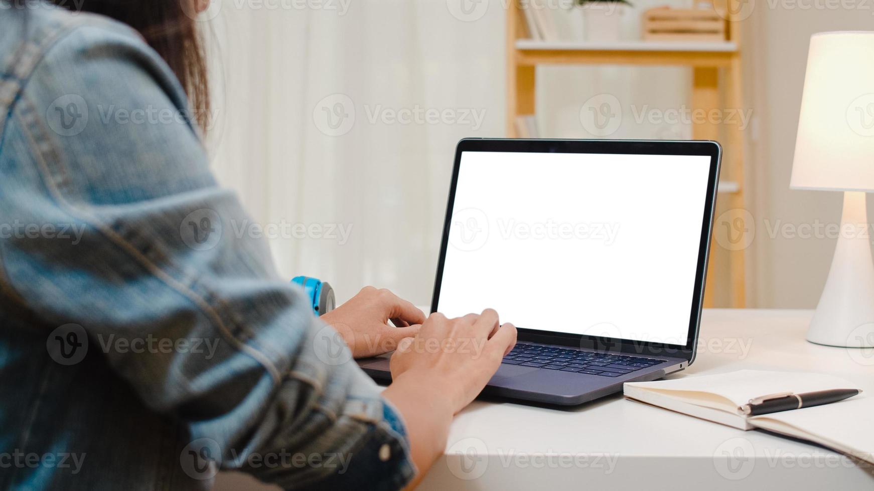 Mujer joven creativa de Asia sentada en su escritorio usando una computadora portátil con una maqueta de pantalla en blanco en la acogedora sala de estar de la casa moderna. foto