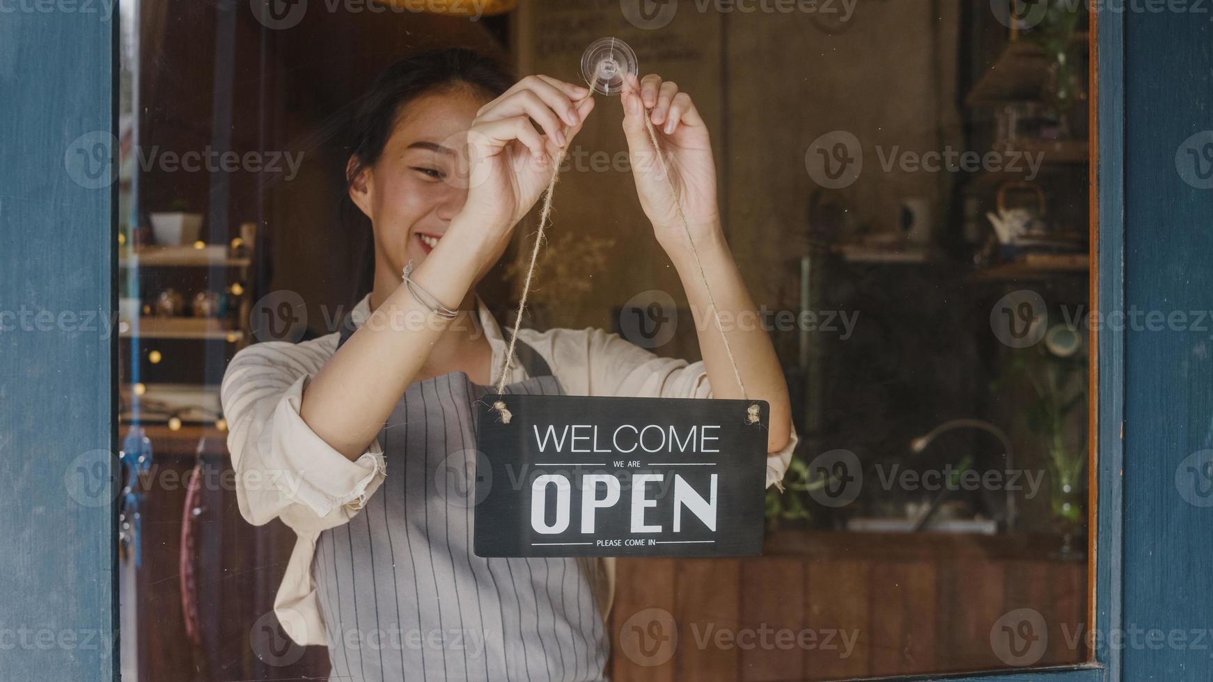 Young Asia manager girl changing a sign from closed to open sign on door cafe looking outside waiting for clients after lockdown. Owner small business, food and drink, business reopen again concept. photo