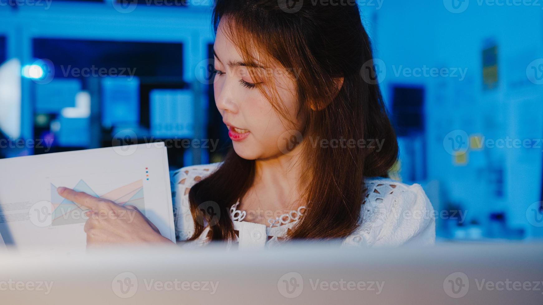 Asia businesswoman social distancing in new normal for virus prevention looking at camera presentation to colleague about plan in video call while work in office night. Lifestyle after coronavirus. photo