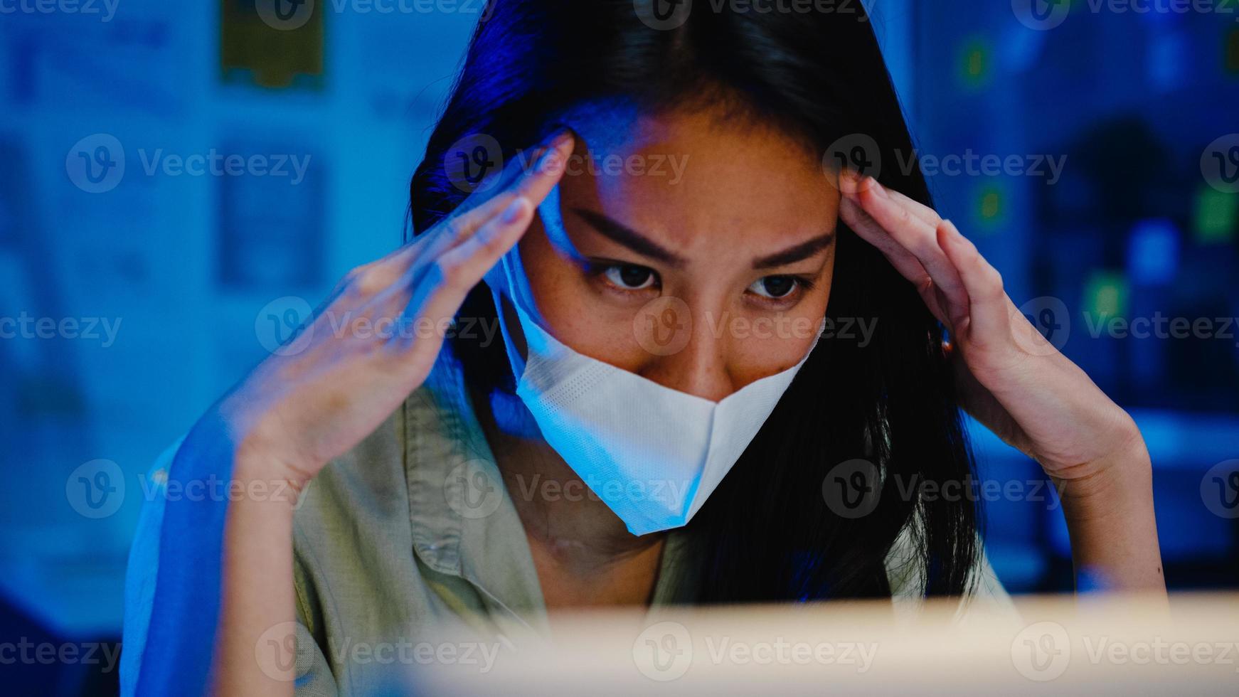 Freelance Asia women wear face mask using laptop hard work at new normal office. Working from home overload at night, self isolation, social distancing, quarantine for corona virus prevention. photo