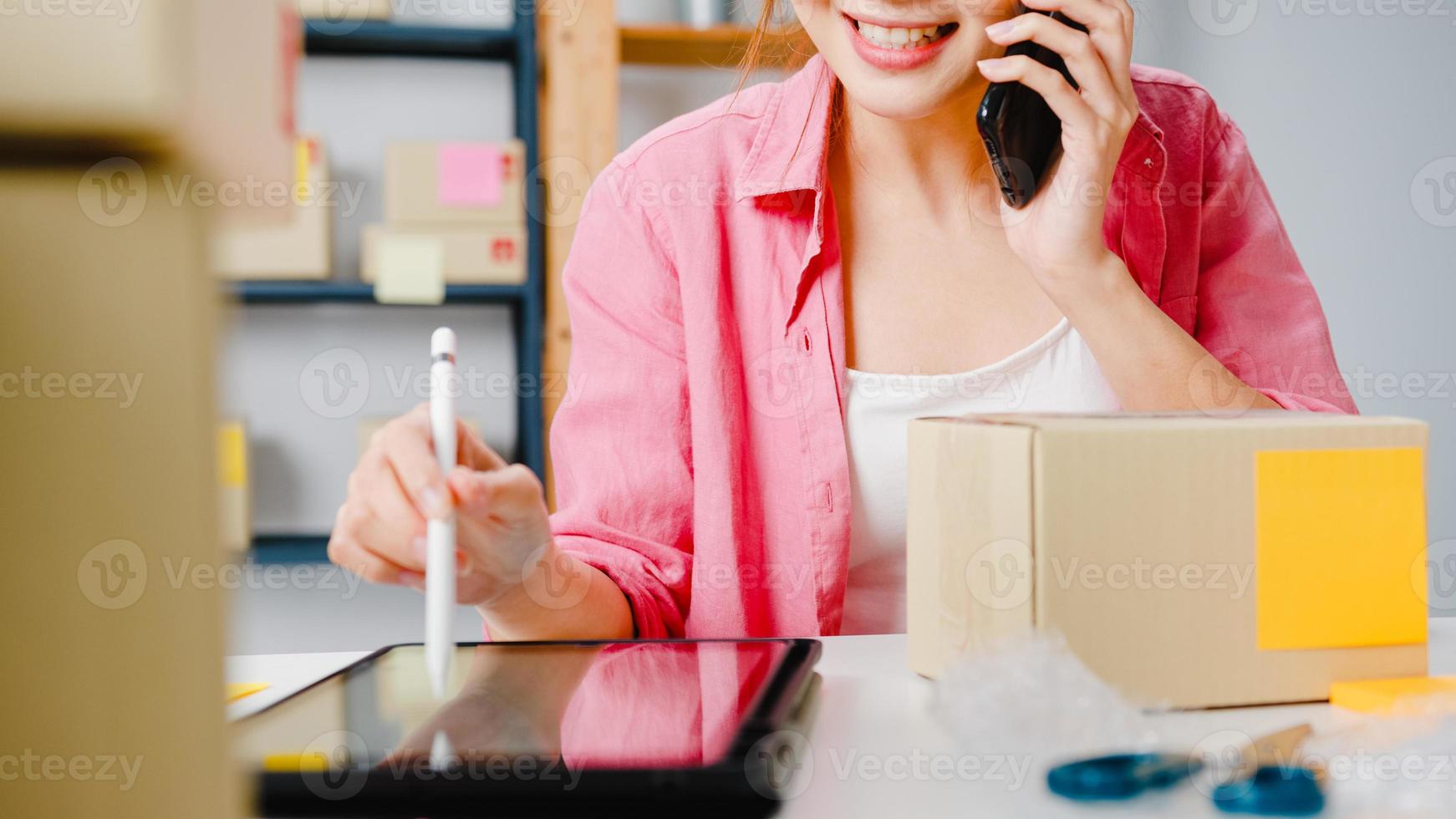 Young Asia businesswoman using mobile phone call receiving purchase order and check product on stock, work at home office. Small business owner, online market delivery, lifestyle freelance concept. photo