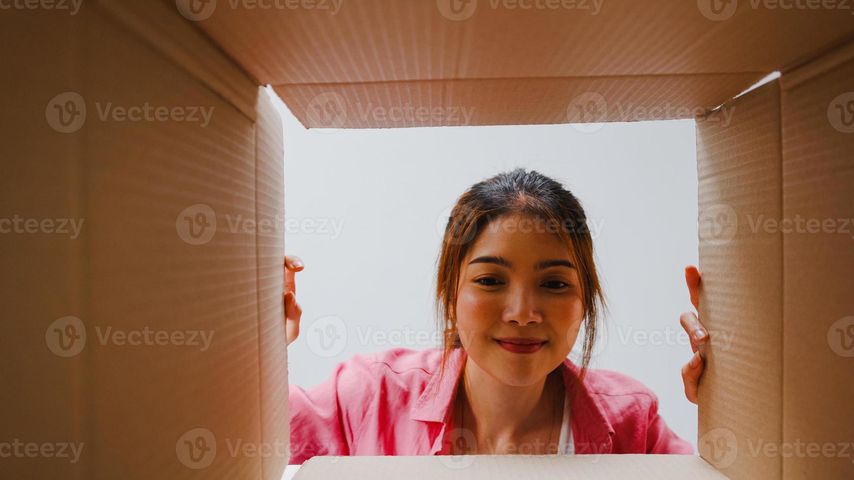 Emocionada joven asiática desempaqueta abriendo una enorme caja de cartón y mirando dentro de un nuevo regalo en casa. feliz clienta millennial satisfecha con la compra ordenada. entrega y concepto de compras en línea. foto