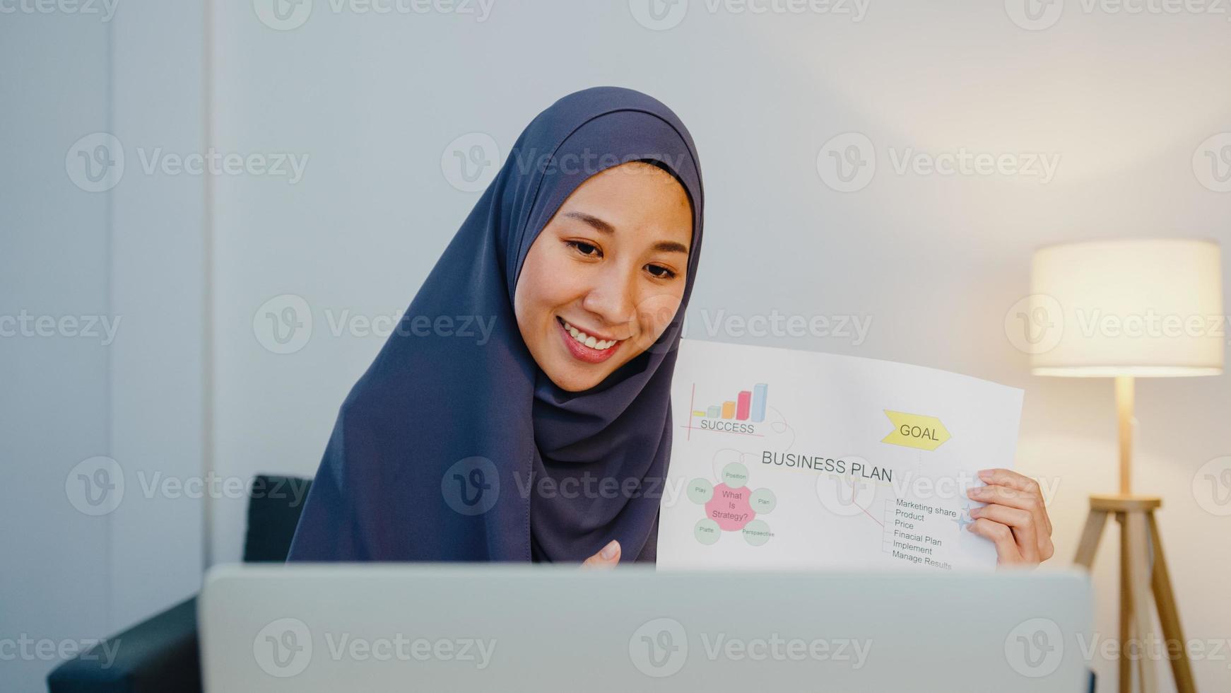 Asia dama musulmana usa hijab usando computadora portátil, hable con su colega sobre el plan en una reunión de videollamada mientras trabaja de forma remota desde casa por la noche en la sala de estar. distanciamiento social, cuarentena por coronavirus. foto