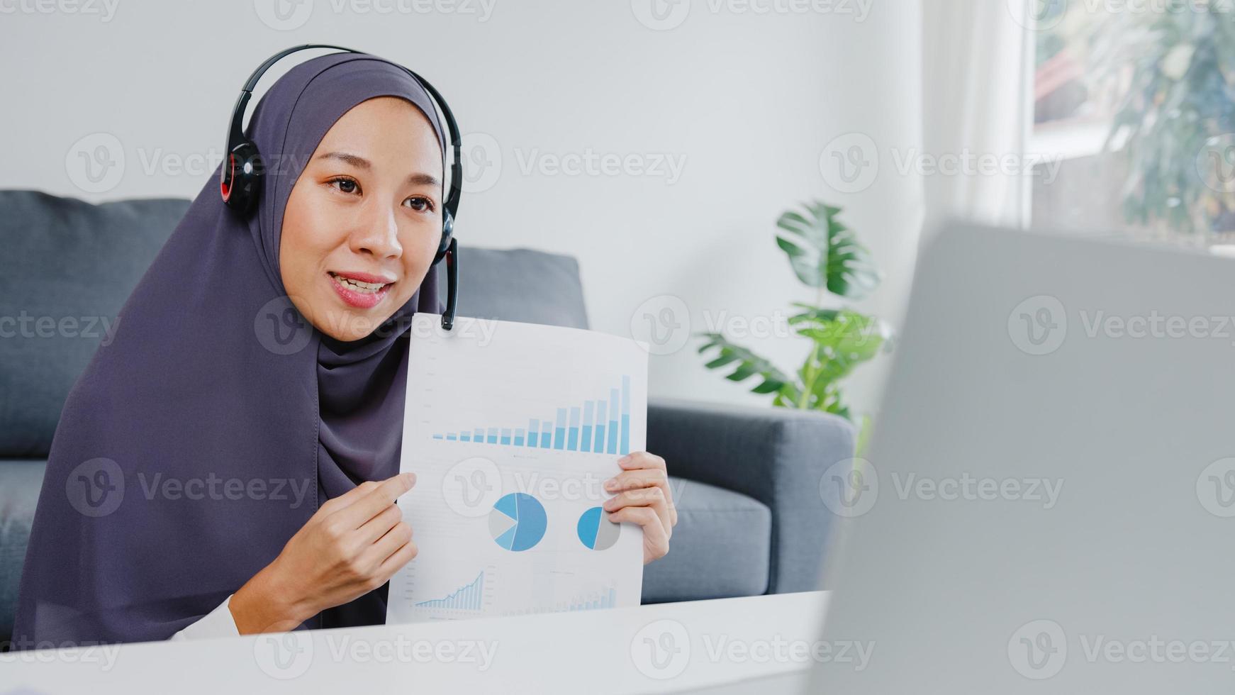 Asia muslim lady wear headphone using laptop talk to colleagues about sale report in conference video call while working from home at living room. Social distancing, quarantine for corona virus. photo