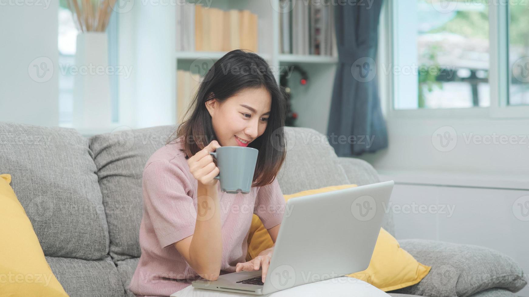 Mujer asiática independiente de negocios joven que trabaja en la computadora portátil revisando las redes sociales y tomando café mientras está acostado en el sofá cuando se relaja en la sala de estar en casa. mujeres de estilo de vida en el concepto de casa. foto