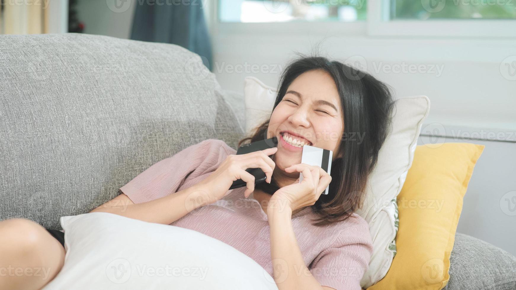 Young smiling Asian woman using smartphone buying online shopping by credit card while lying on sofa when relax in living room at home. Lifestyle latin and hispanic ethnicity women at house concept. photo